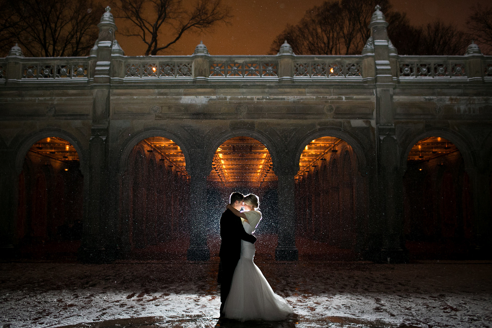 A couple in wedding attire kissing under a bridge at night, with water splashing around them, illuminated by warm lights.