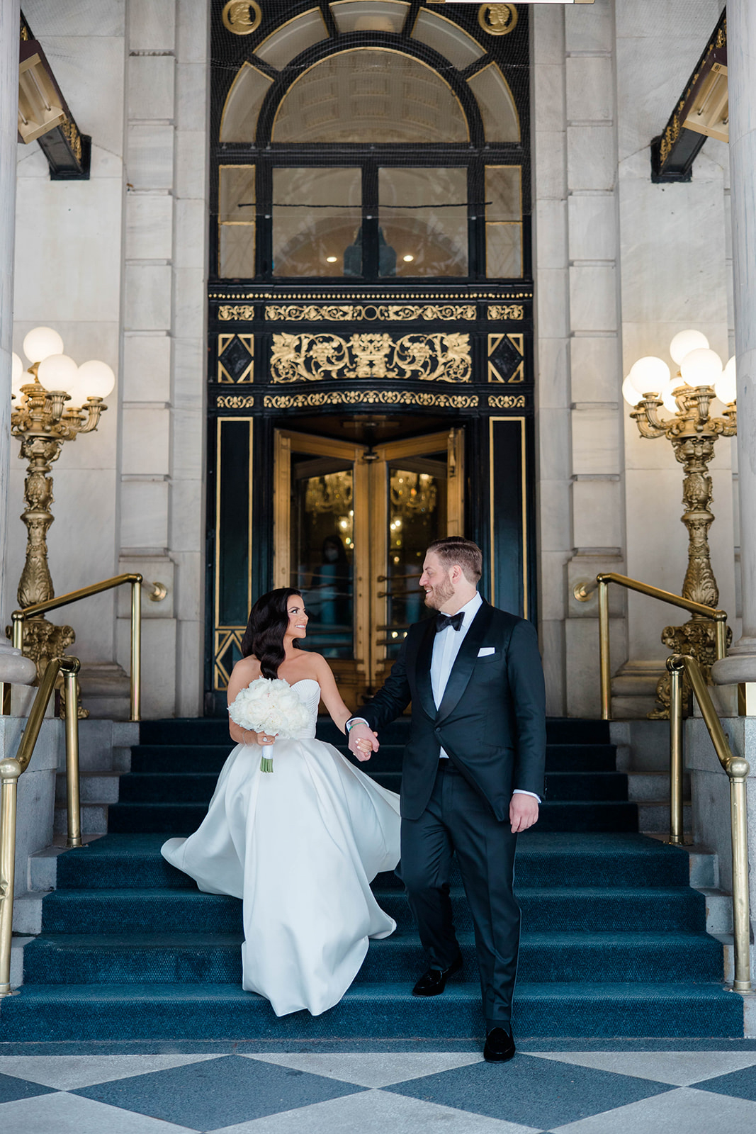 Bride and groom holding hands on stairs in front of grand entrance, ornate gold and black details, elegant lighting fixtures.