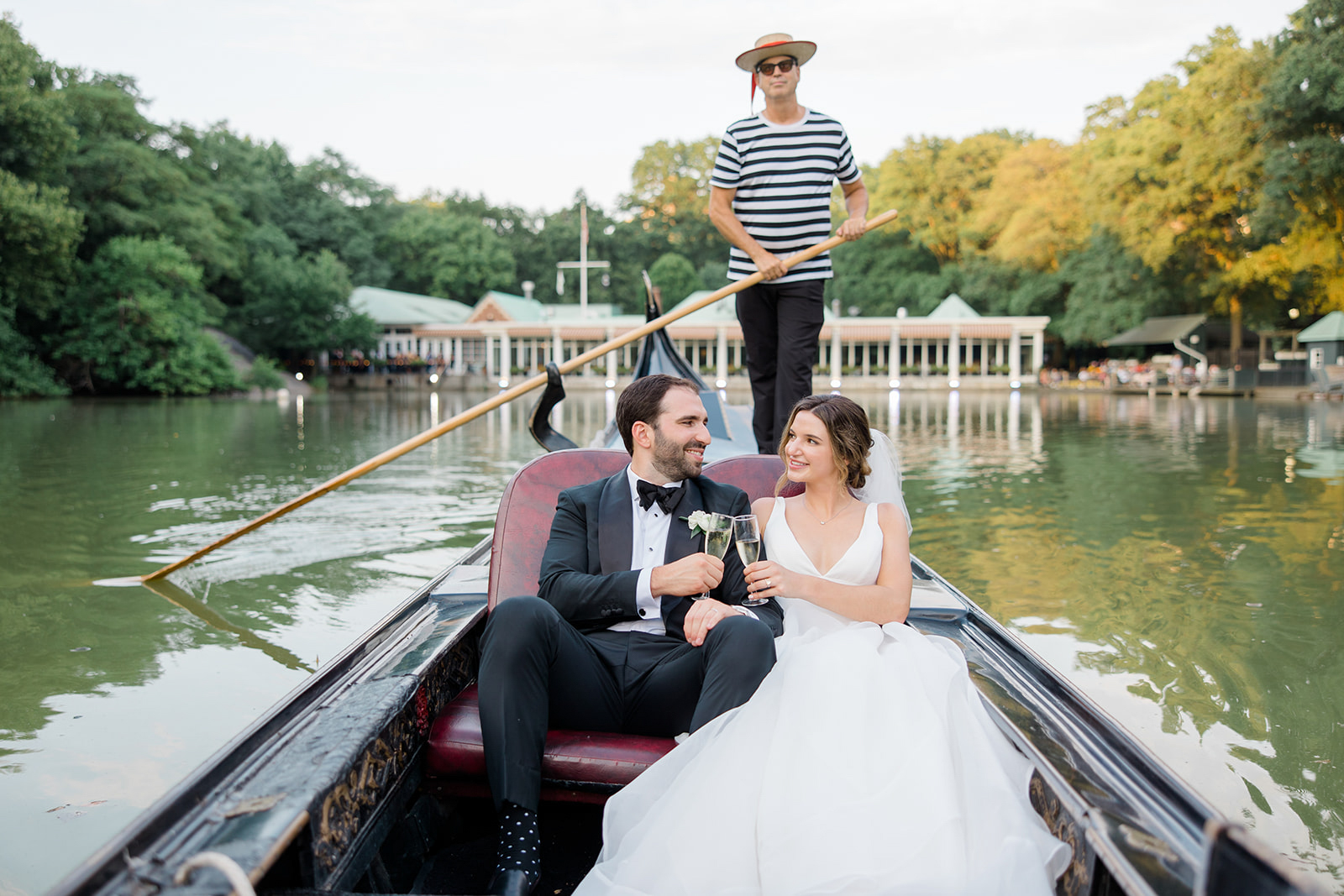A couple in wedding attire sitting in a boat on a river, with a person standing on a boat behind them, holding a pole. Trees and houses are in the background.