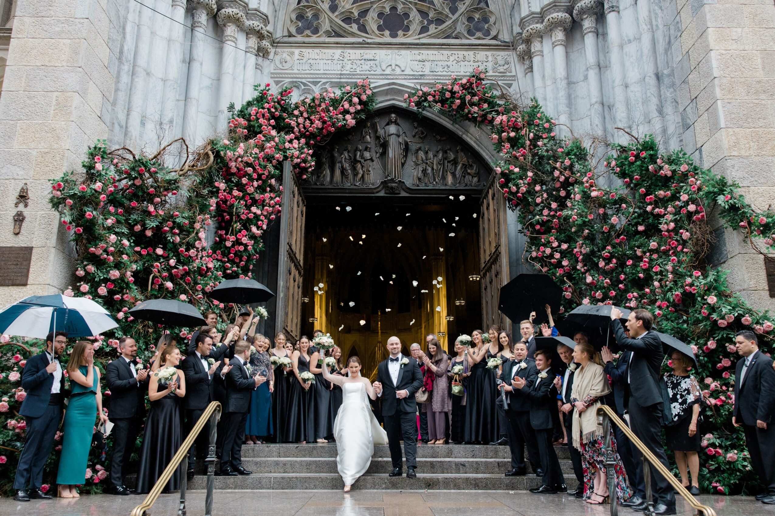 Bride and groom walking down steps outside a decorated church, surrounded by guests holding umbrellas, floral archway above.