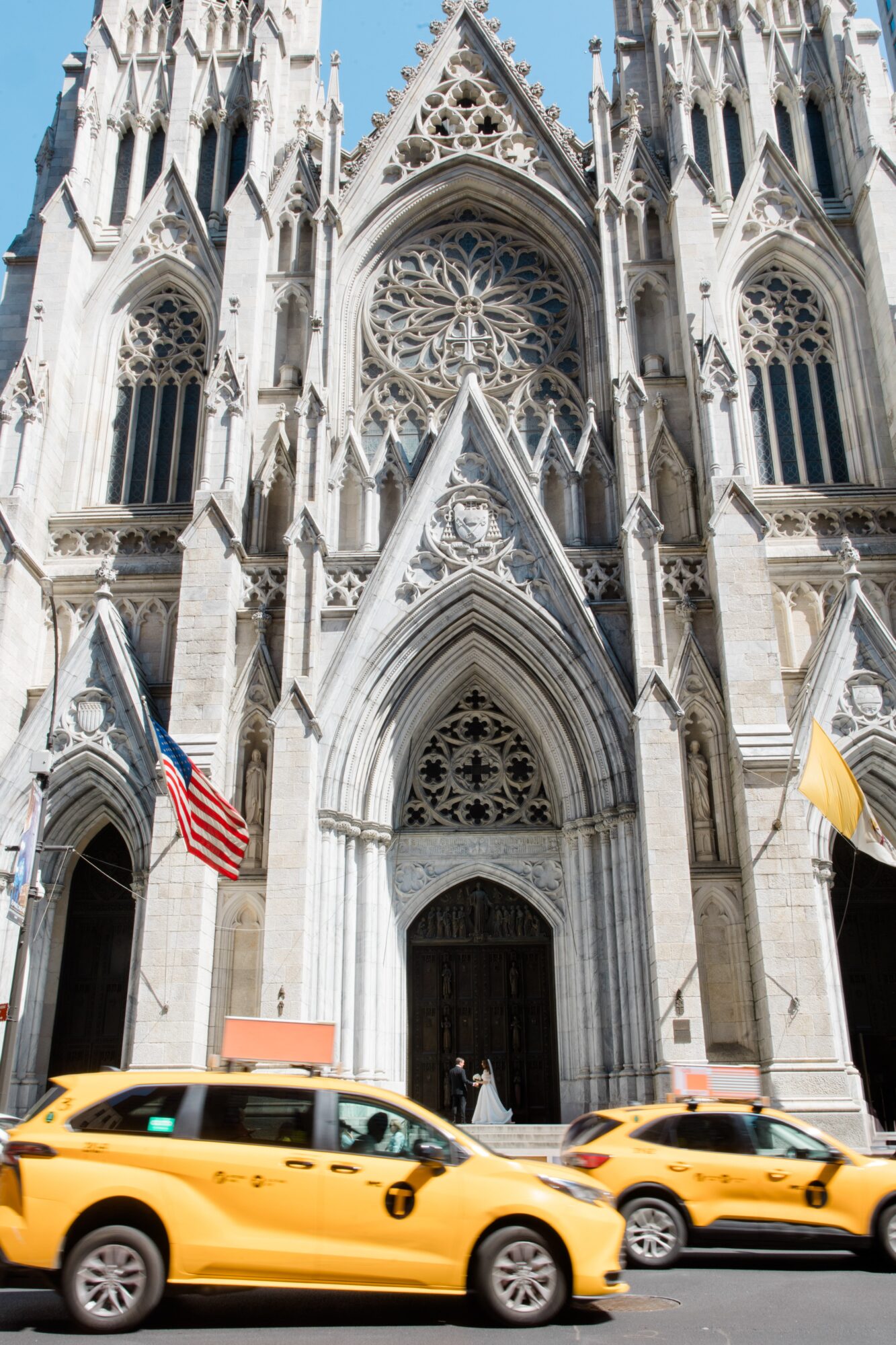 Facade of a Gothic cathedral with detailed architecture, flags, and yellow taxis in front.
