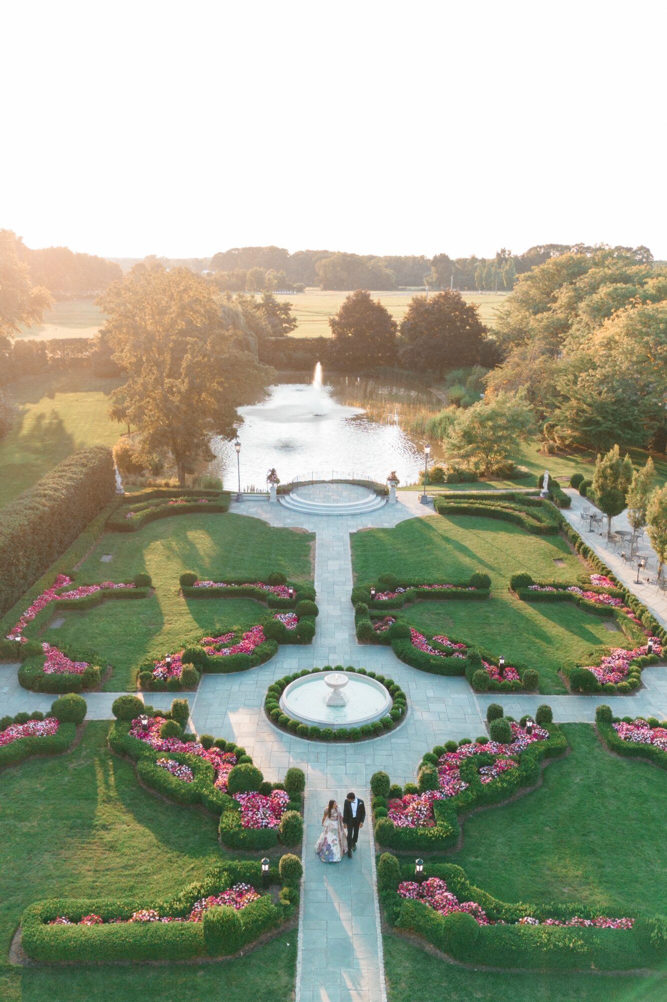 A garden with fountains, flower beds, and pathways viewed from above, with trees and a water feature in the background.