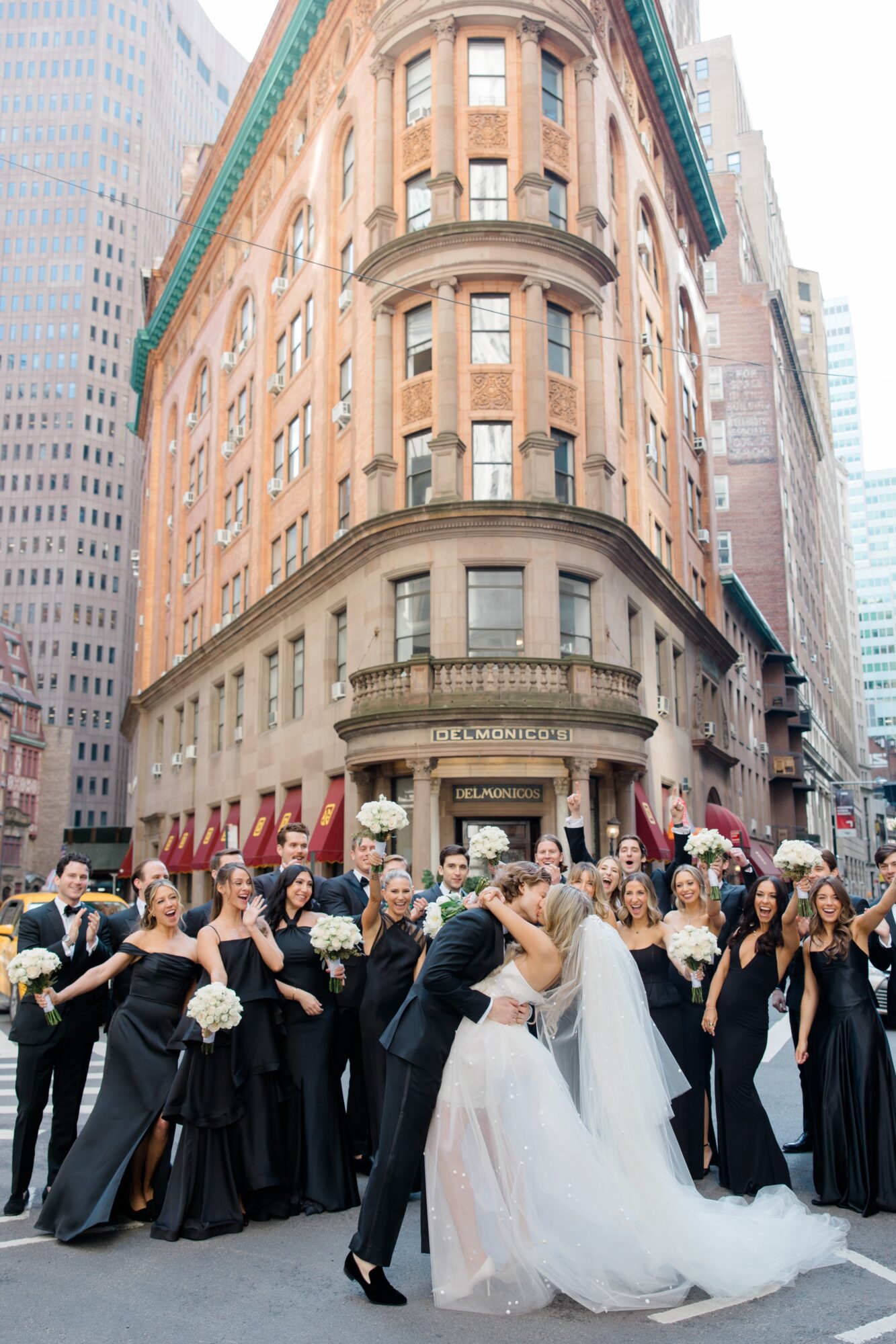 Wedding couple kissing surrounded by bridesmaids and guests on city street, tall building in background.