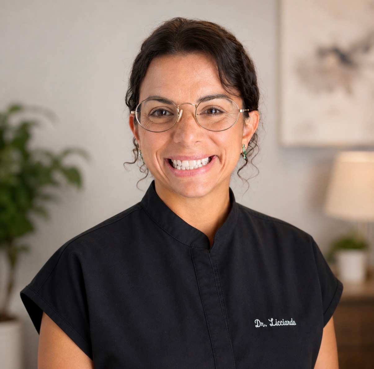 Smiling woman with glasses and dark hair, wearing a black uniform with embroidered name, in a room with a plant and lamp.