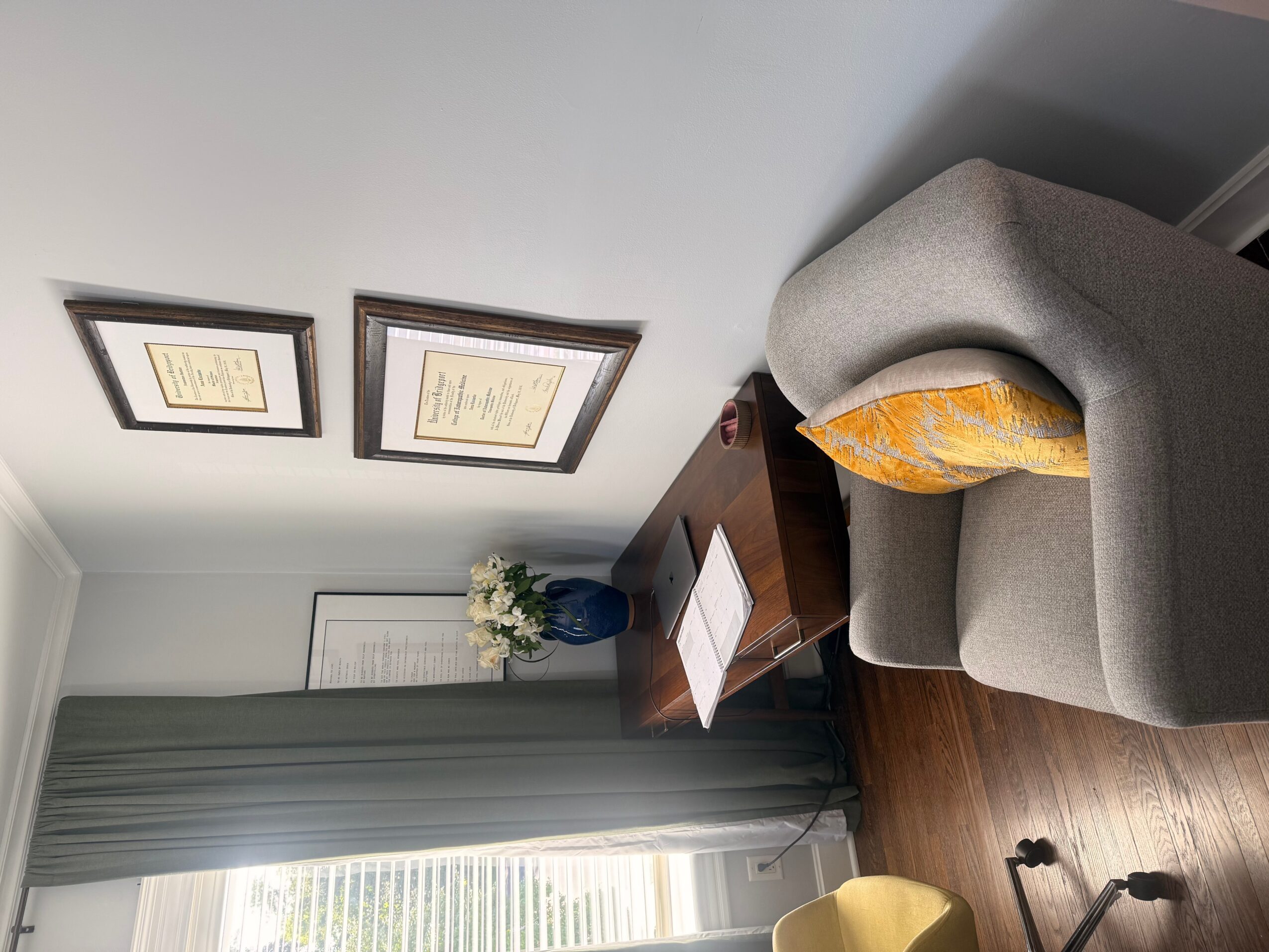 Office corner with framed certificates, a gray armchair, a wooden side table with a document, and a window with curtains.