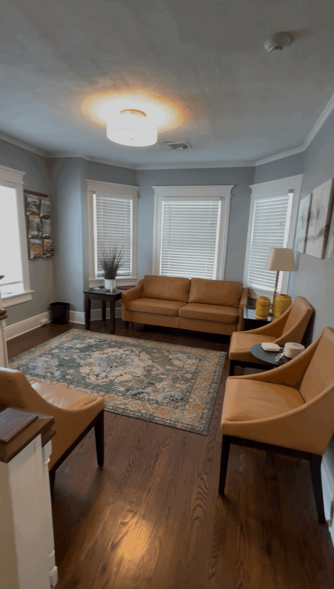 Living room with three windows, a tan sofa, two yellow chairs, a floor lamp, and a patterned rug.