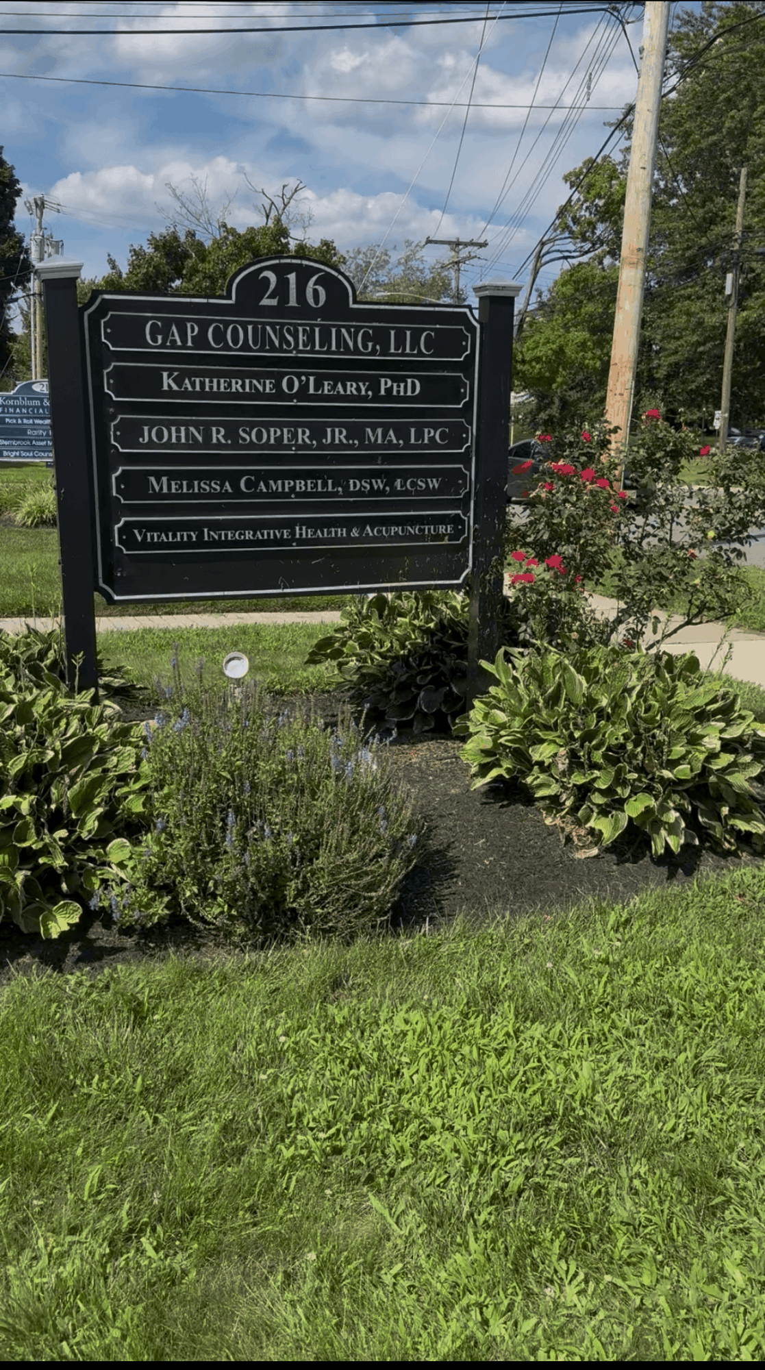 Black and white sign with multiple names and titles, surrounded by green plants and bushes, under a partly cloudy sky.