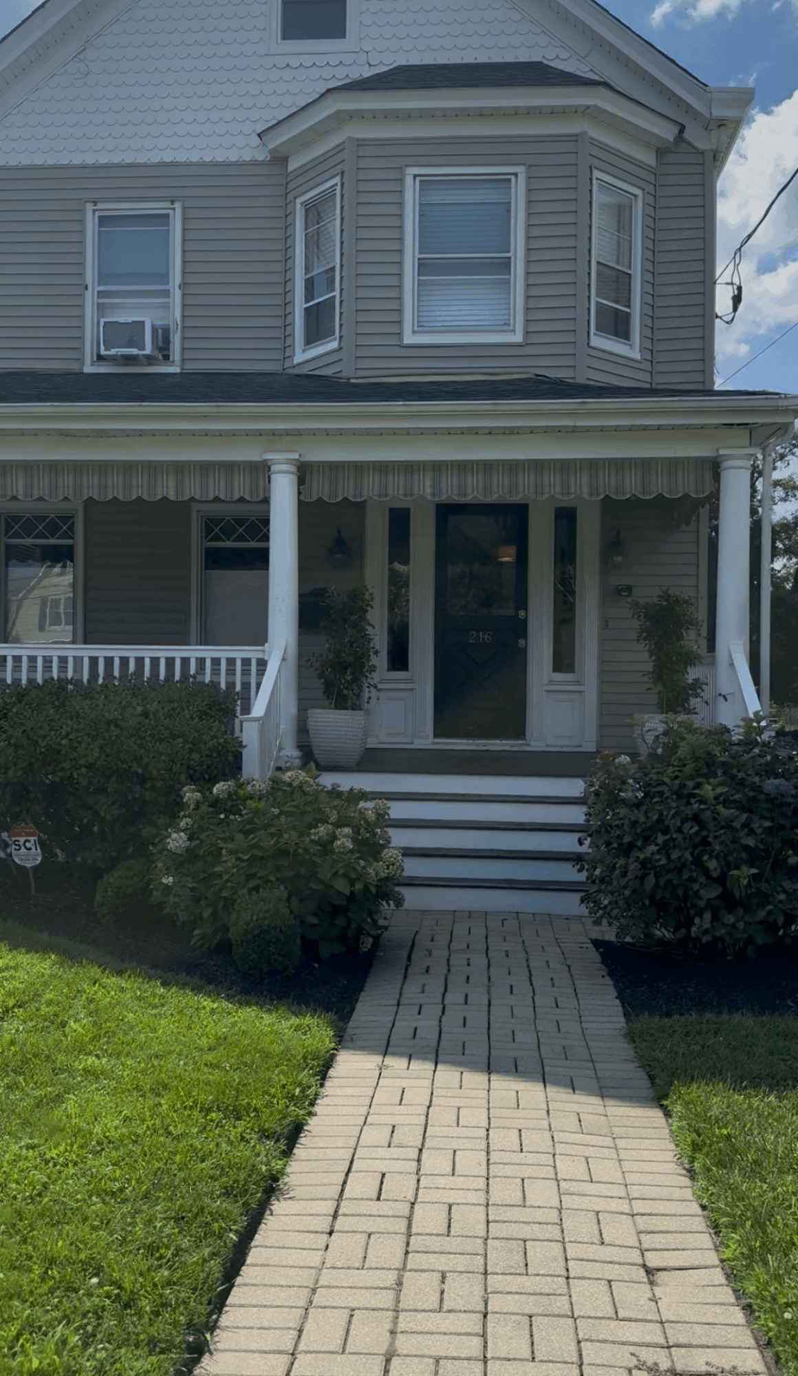 Front view of a house with a porch, steps, and a brick walkway leading to the entrance, surrounded by greenery.