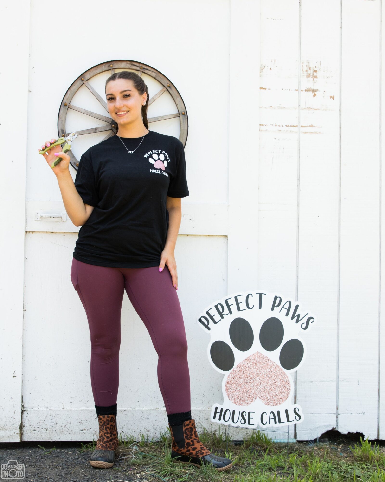 Young woman standing outdoors near a white wall, holding a small plant, smiling, wearing a black T-shirt and maroon pants.