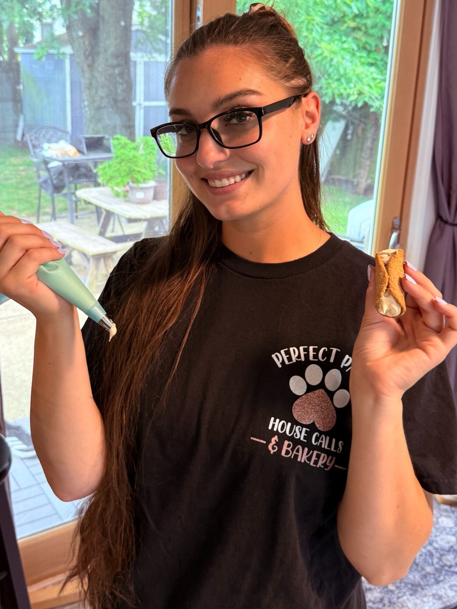 Young woman with glasses smiling, holding a piping bag and a cookie, standing indoors near a window with a garden view.
