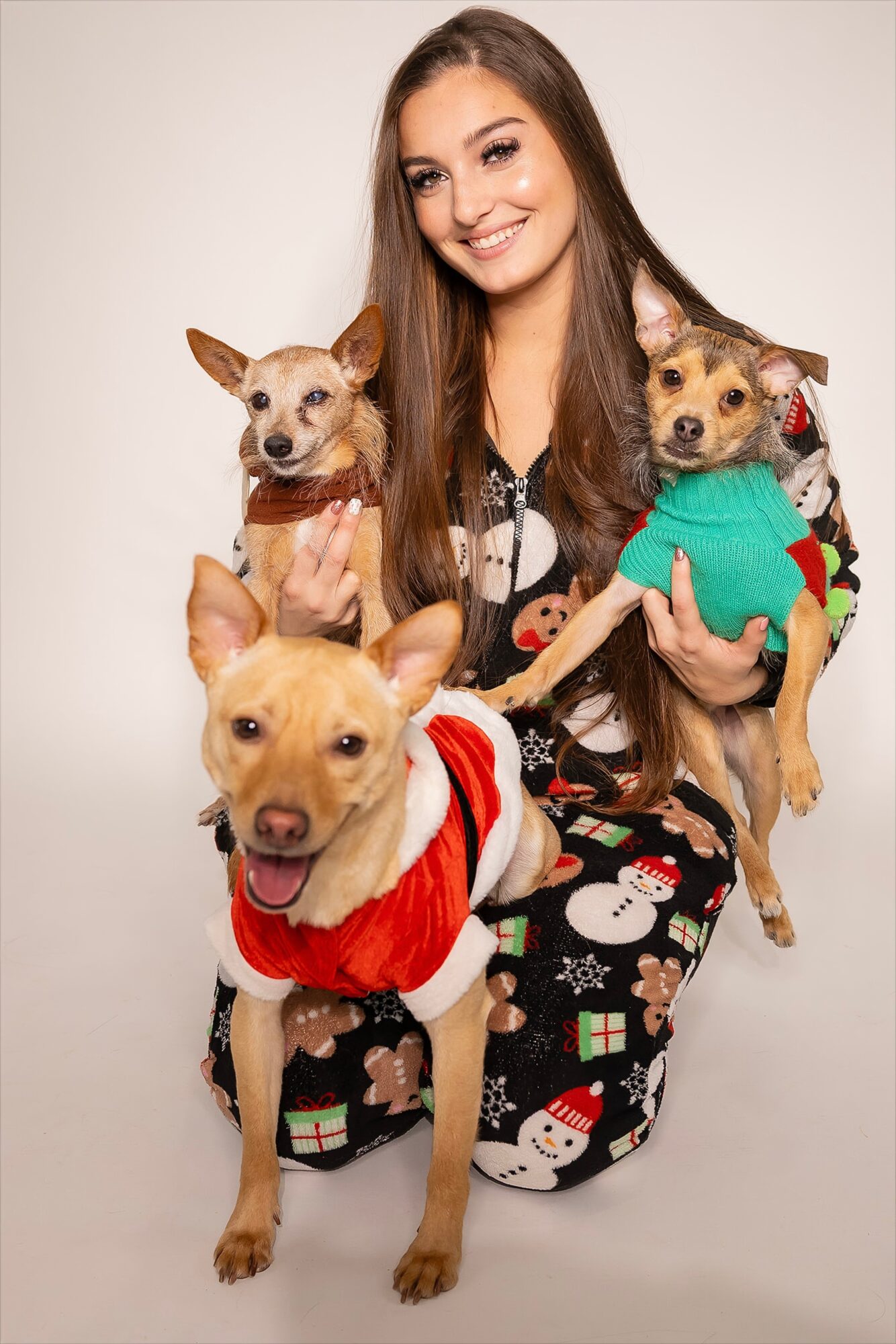 Young woman with long brown hair smiling, holding three small dogs dressed in festive clothing, against a plain background.