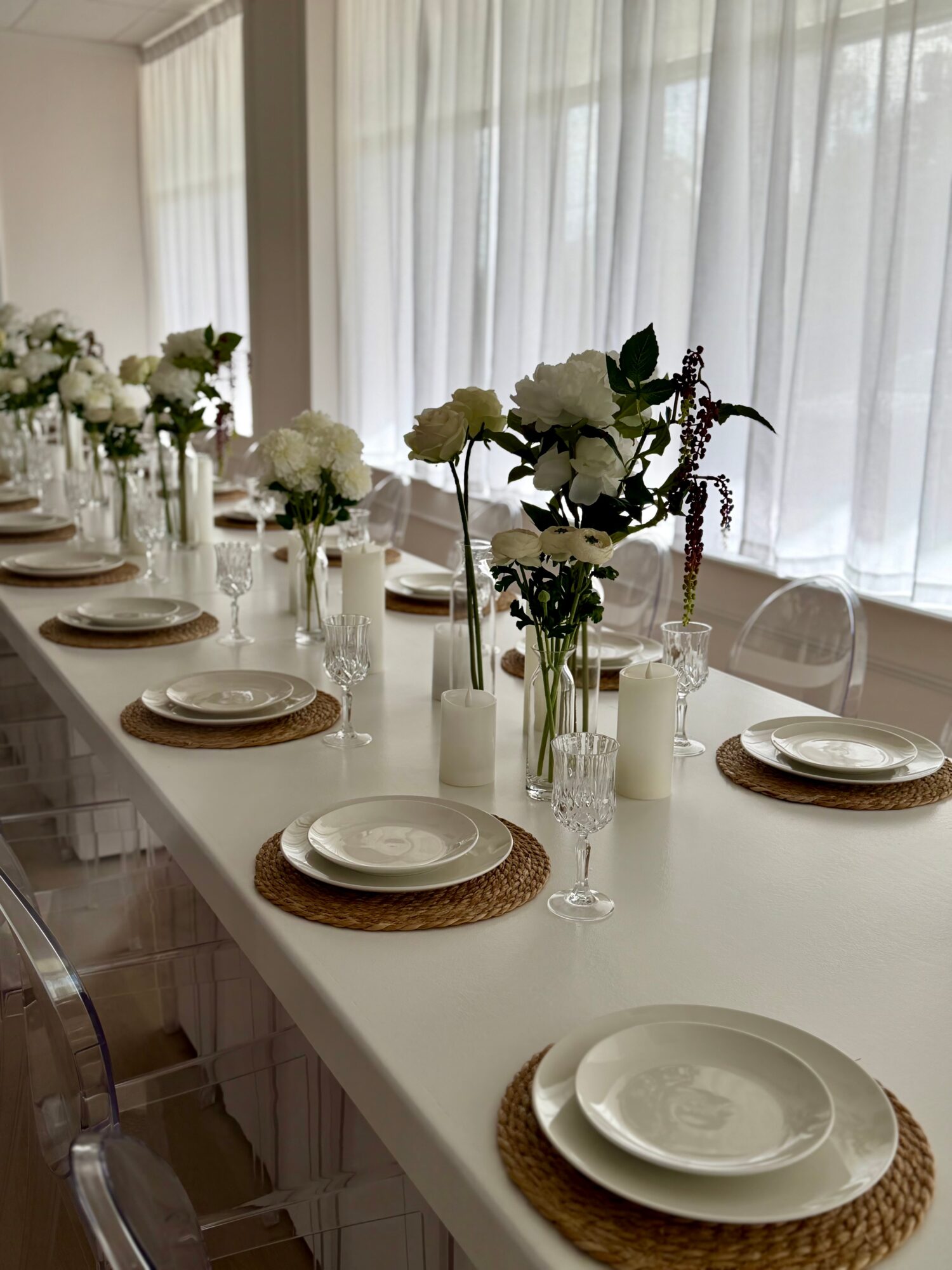 Table set with white plates, glasses, and floral centerpieces in vases, near large windows with sheer curtains