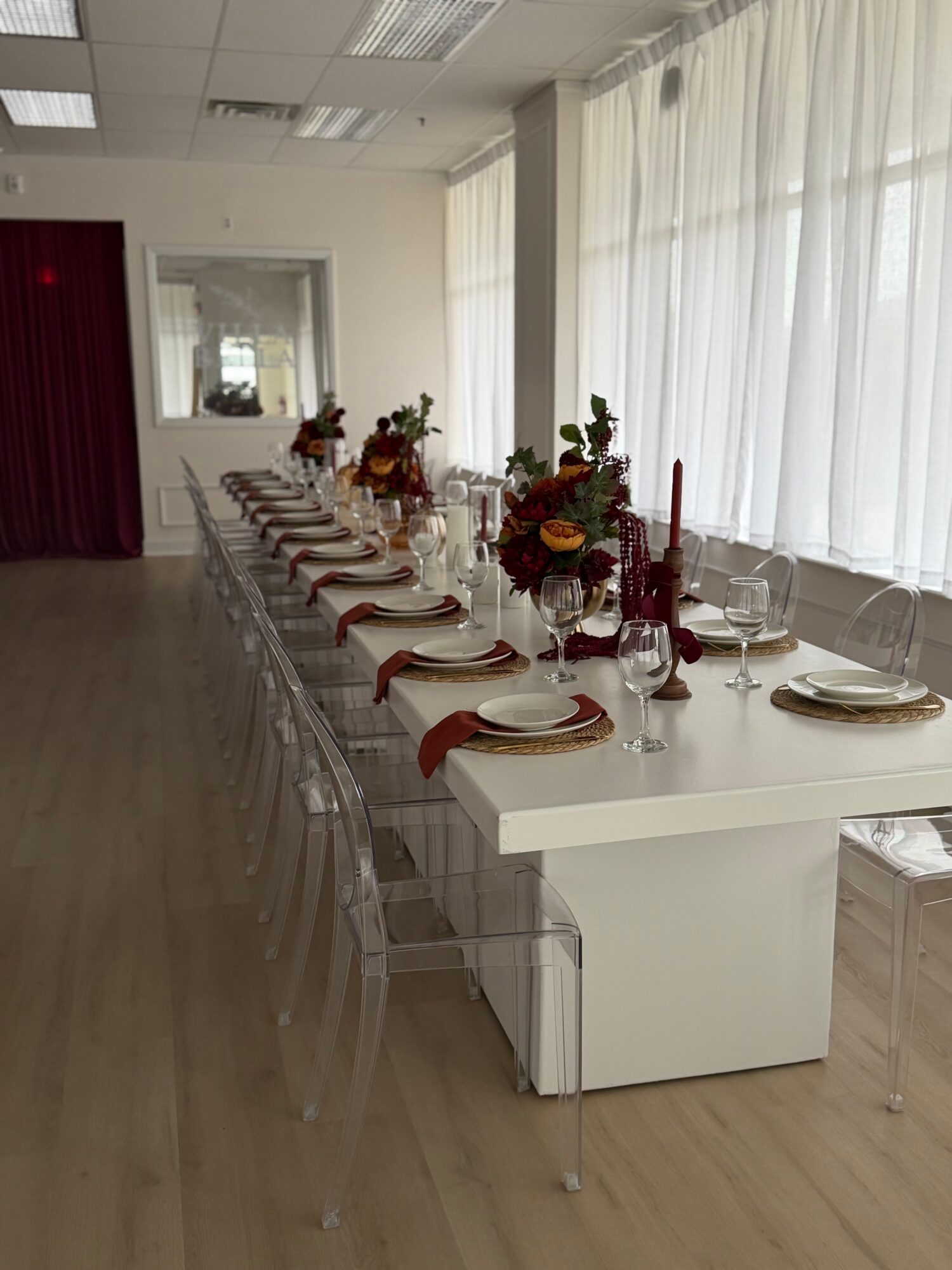 Long dining table set with plates, glasses, and floral centerpieces in a bright room with large windows and white curtains.