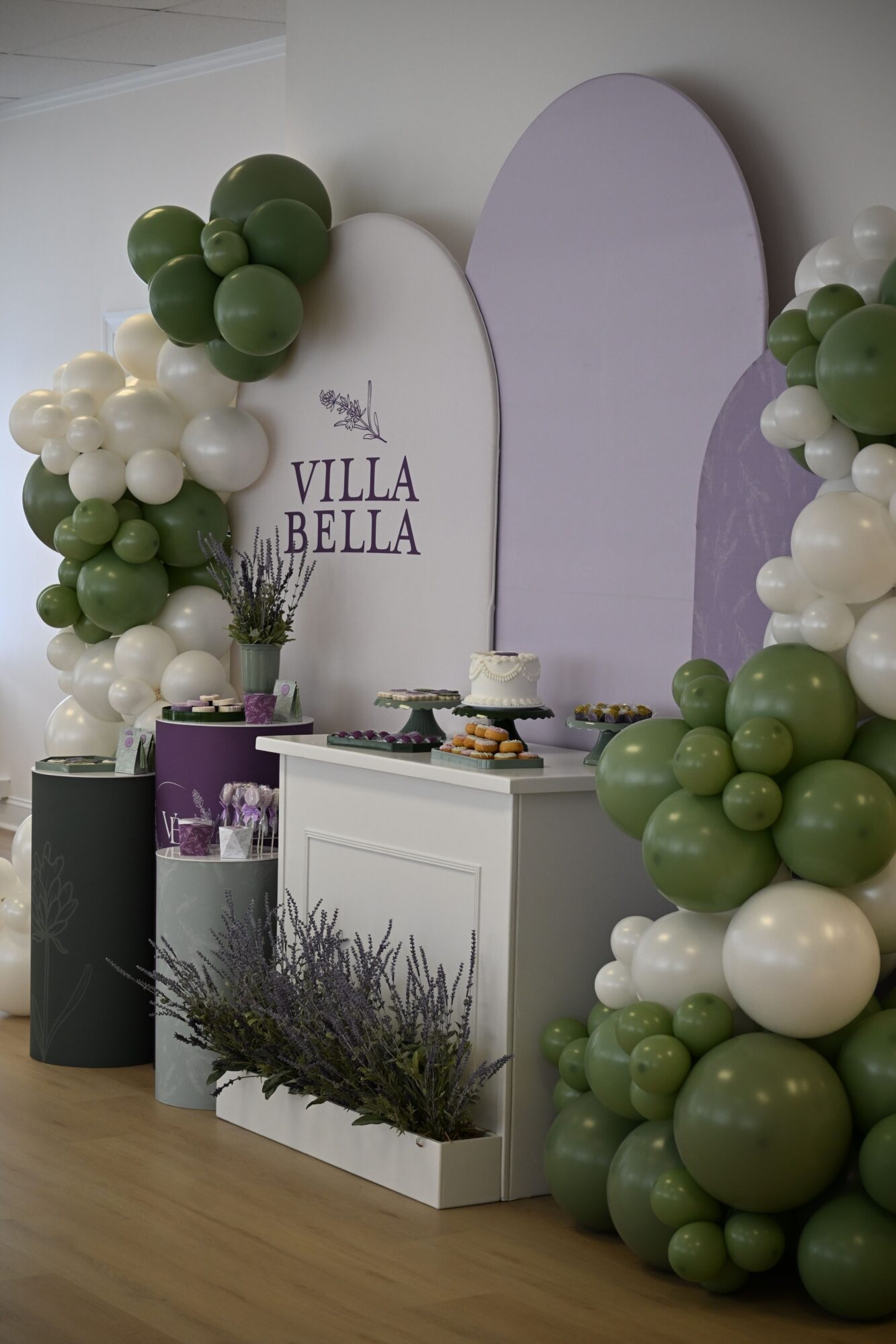 Decorative arrangement with green, white, and purple balloons, candles, and a lavender sprig on a table, with a white and purple backdrop.