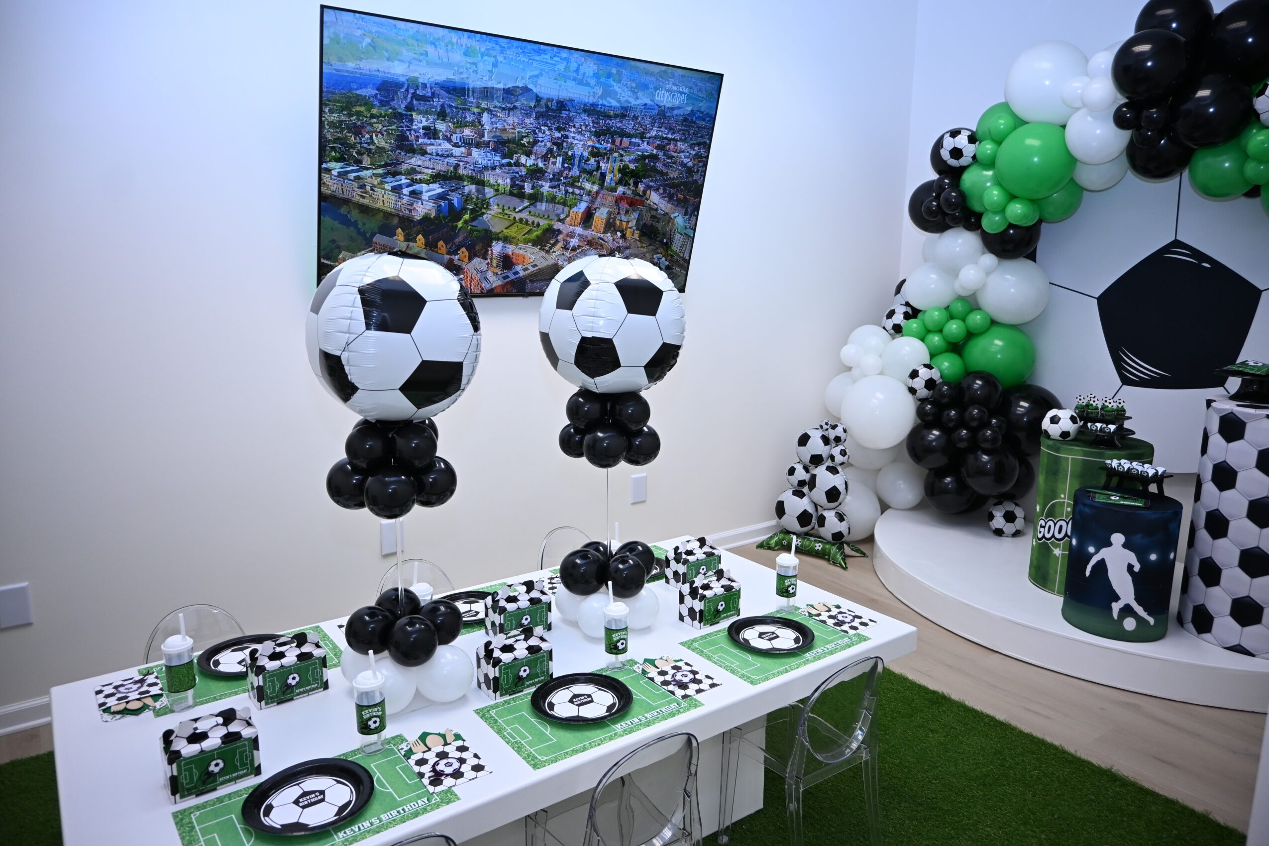 Party setup with soccer-themed decorations, balloons, and a large screen displaying a cityscape, in a white room.