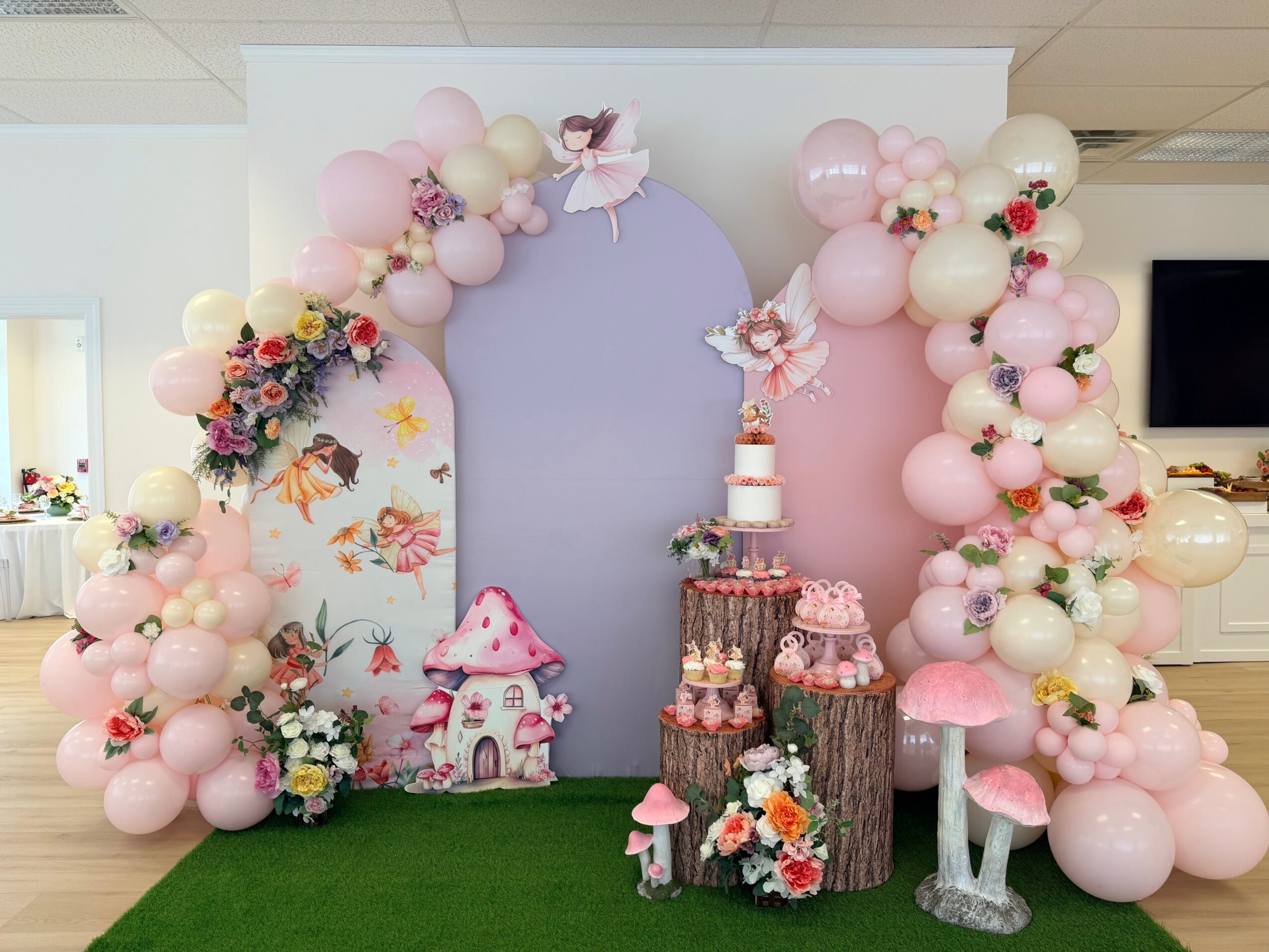 Decorative arch with pink, white, and cream balloons, flowers, and fairy-tale themed elements, including a mushroom house and a cake display.