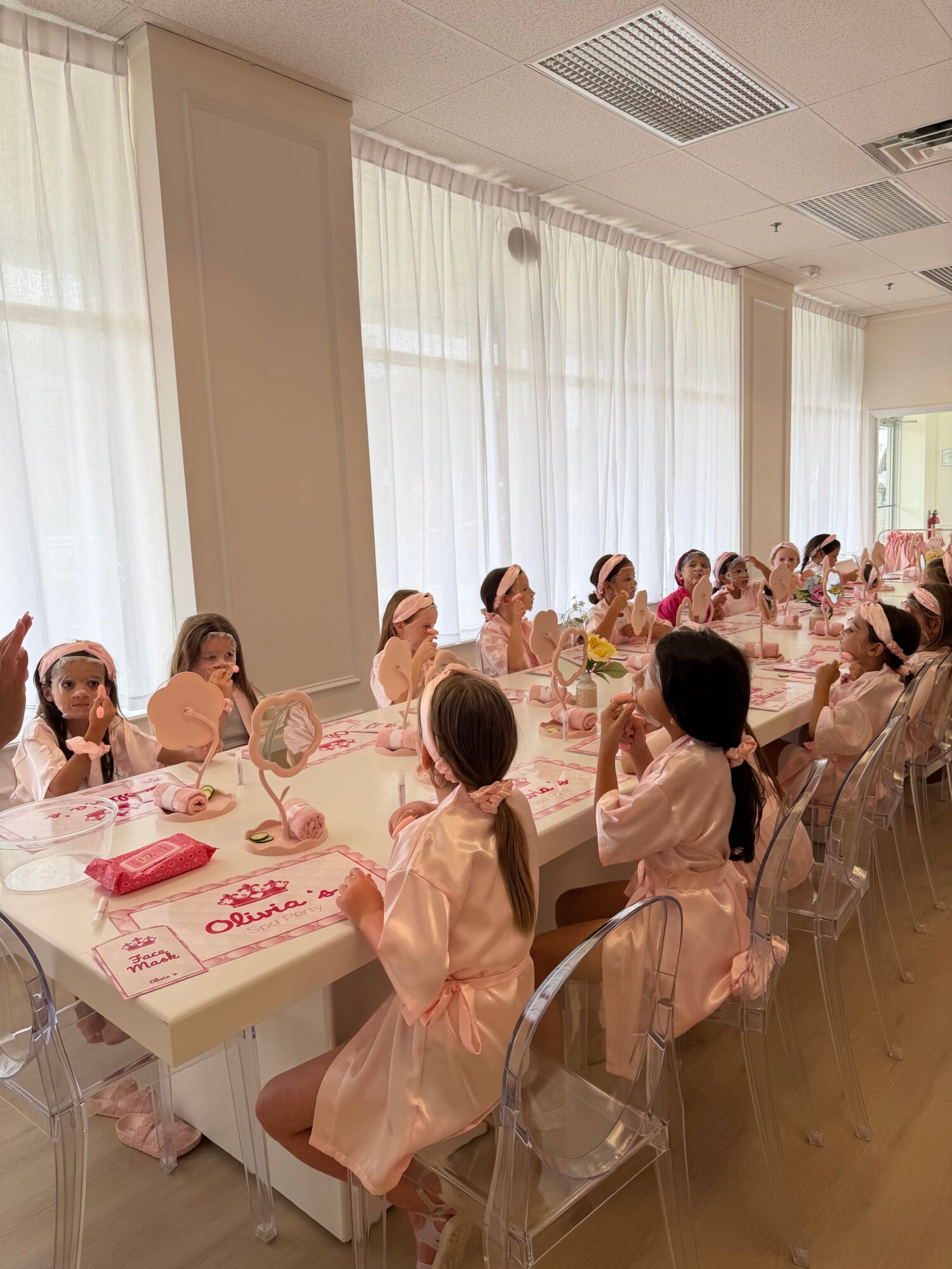 People in pink dresses seated at a long table during a celebration or event, with a window and curtains in the background.