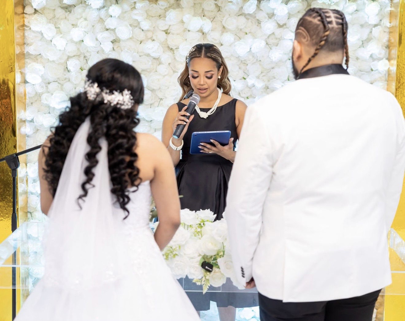 A couple faces an officiant holding a book amid white floral decorations.