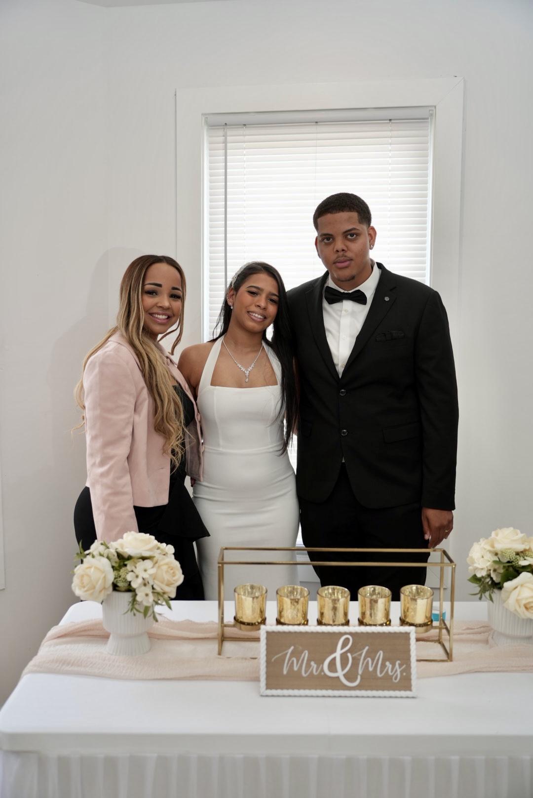 Three people pose behind a decorated table with flowers, candles, and a sign in front of a window.