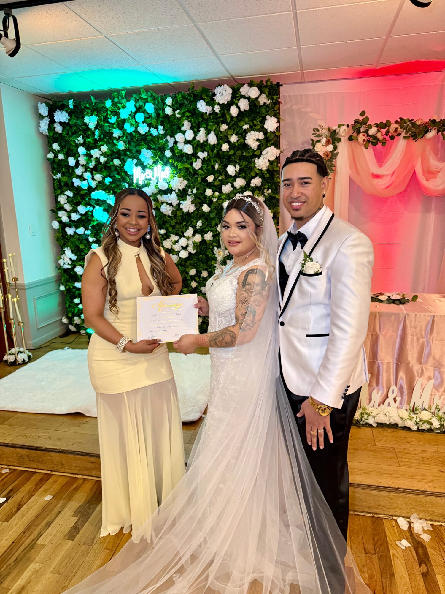 Three people pose together at a wedding reception in front of a flower wall.