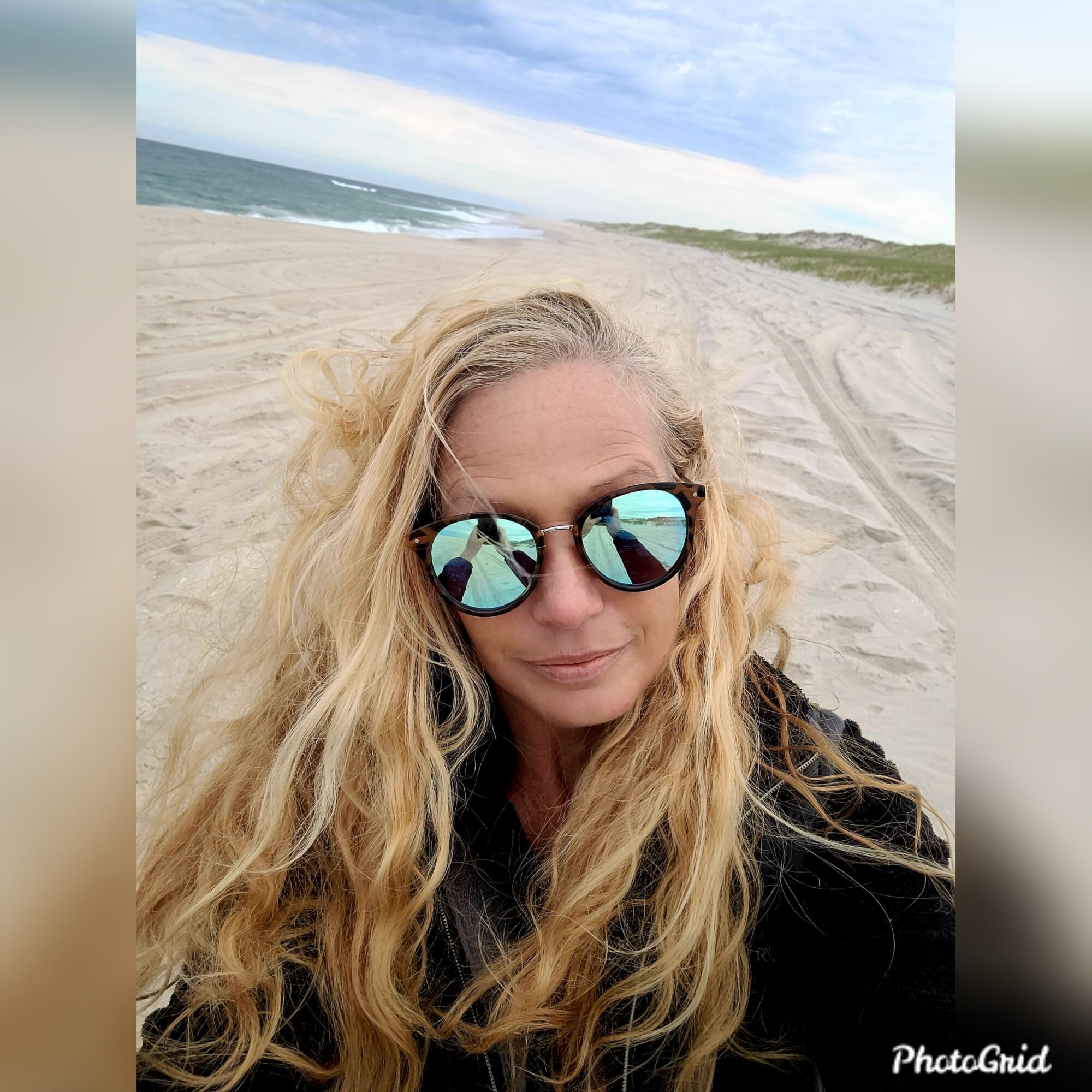 A person wearing reflective sunglasses takes a selfie on a sandy beach, with the ocean and cloudy sky behind them.