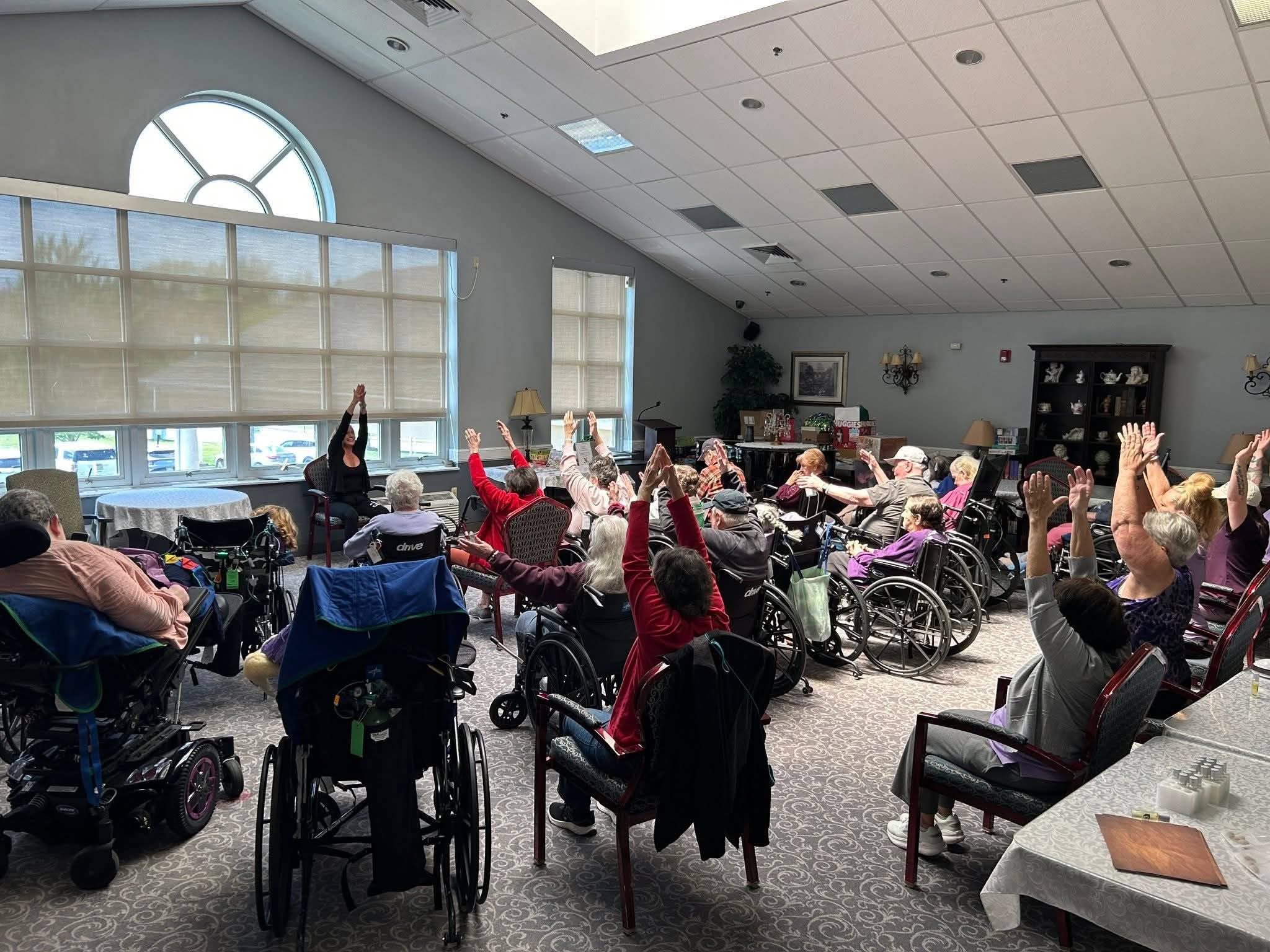 People seated in wheelchairs and chairs raise their hands in a bright community room.