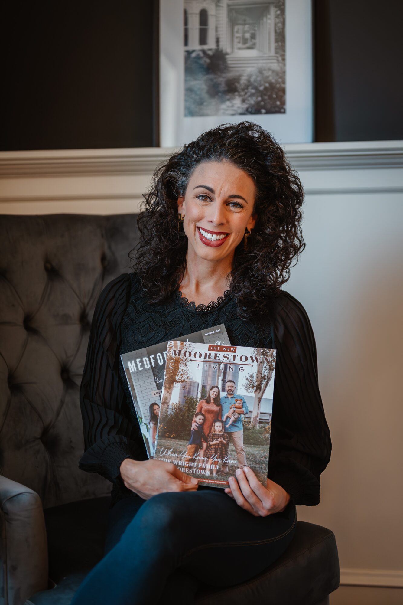 A seated person smiles while holding several magazines in front of a tufted chair.
