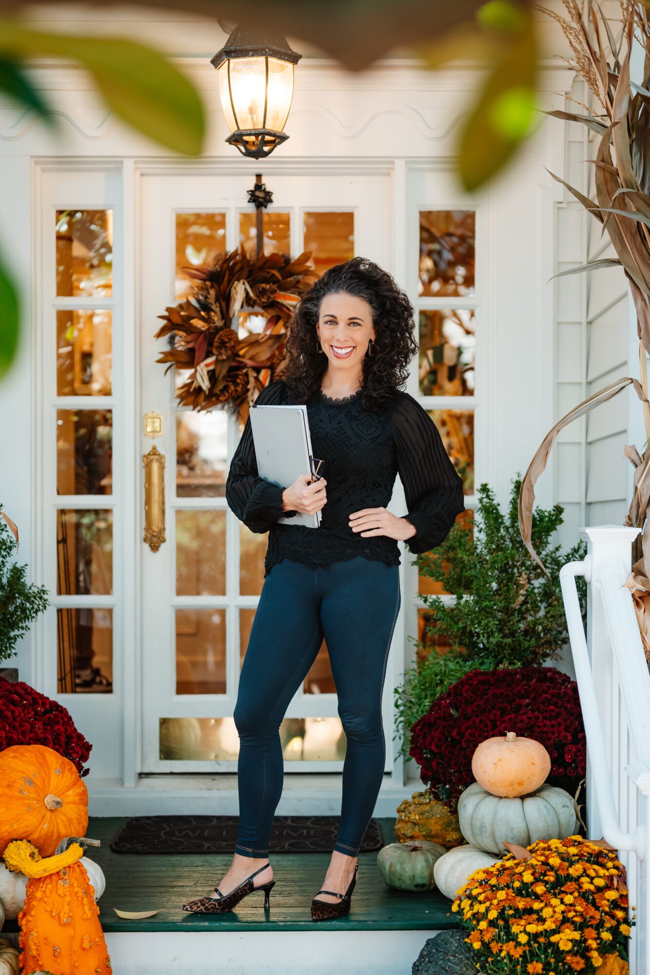 Person holding a tablet stands on a porch beside pumpkins, chrysanthemums, and autumn decorations.