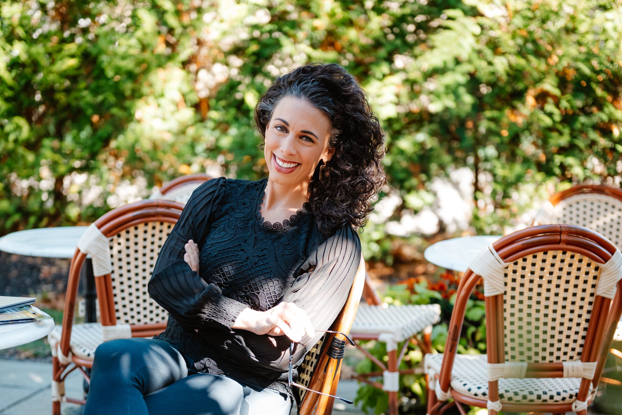 A seated person in a dark top sits among woven café chairs beneath leafy trees.