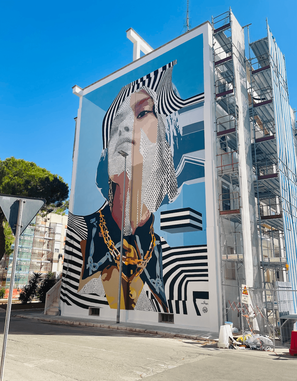 Large mural of a face with striped patterns on a building beside metal scaffolding.