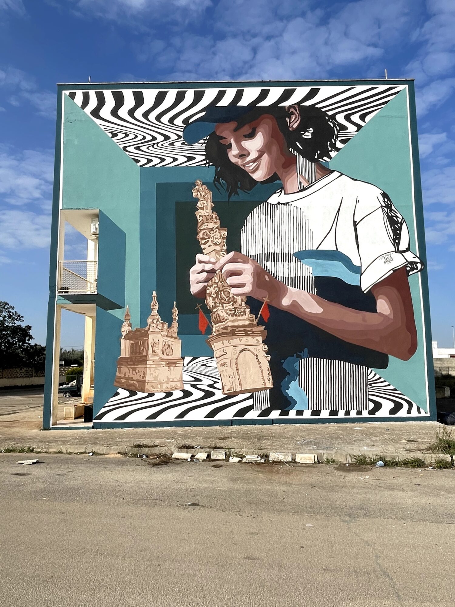 Large outdoor mural depicts a person holding a carved sculpture, framed by black-and-white patterns and a teal background.
