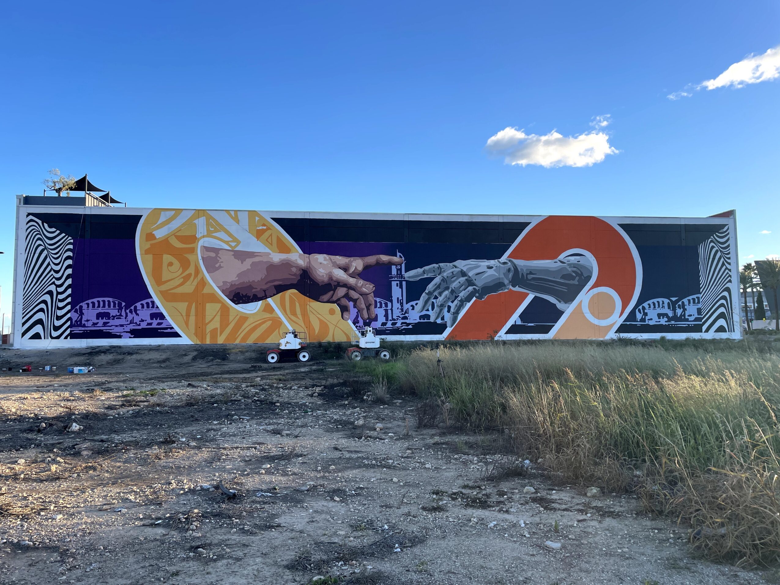 Long mural along a wall depicts an outstretched hand, abstract shapes, and stylized numbers beneath a blue sky.