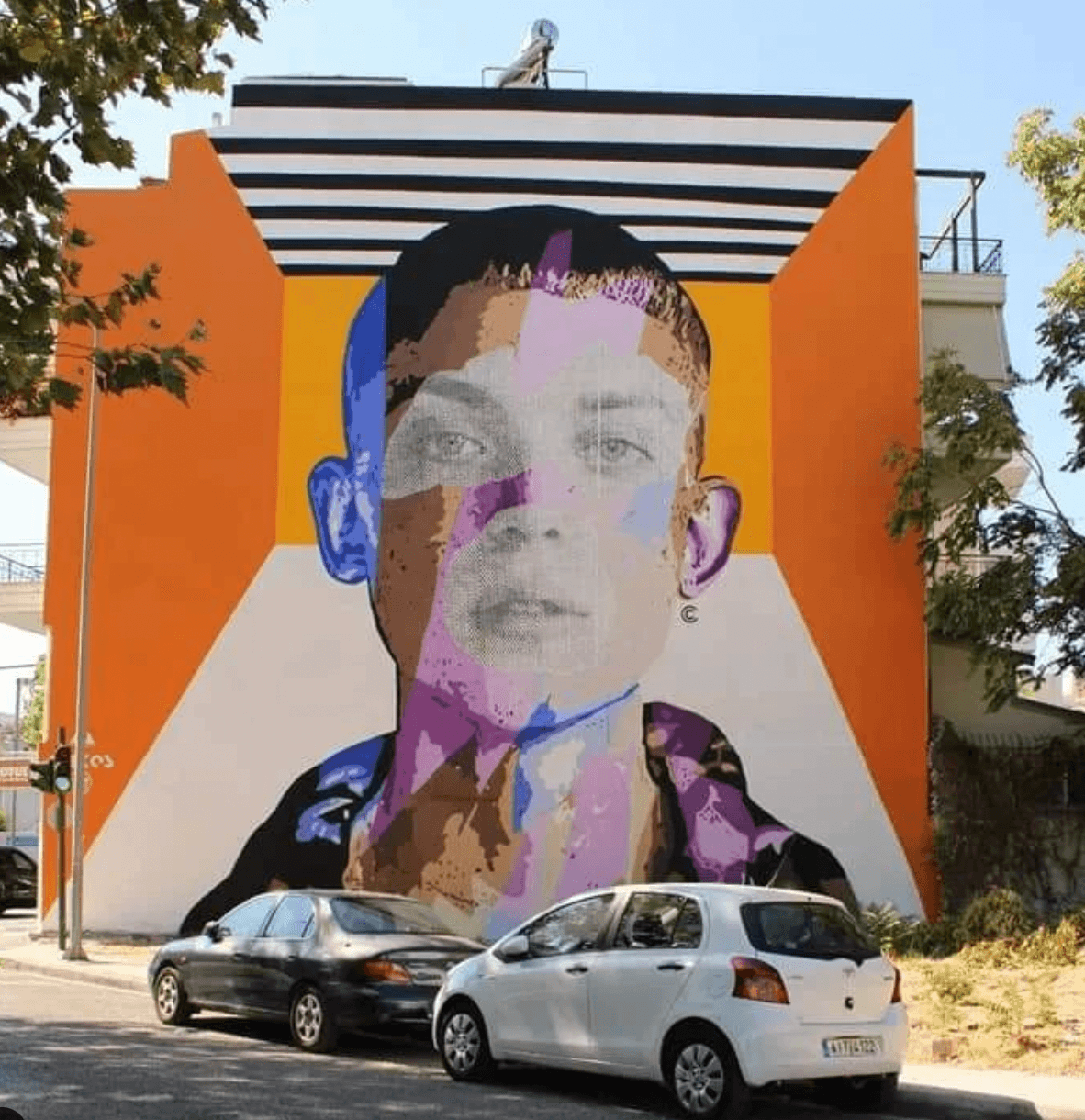 Large colorful portrait mural on an orange building, with two parked cars in front and trees nearby.