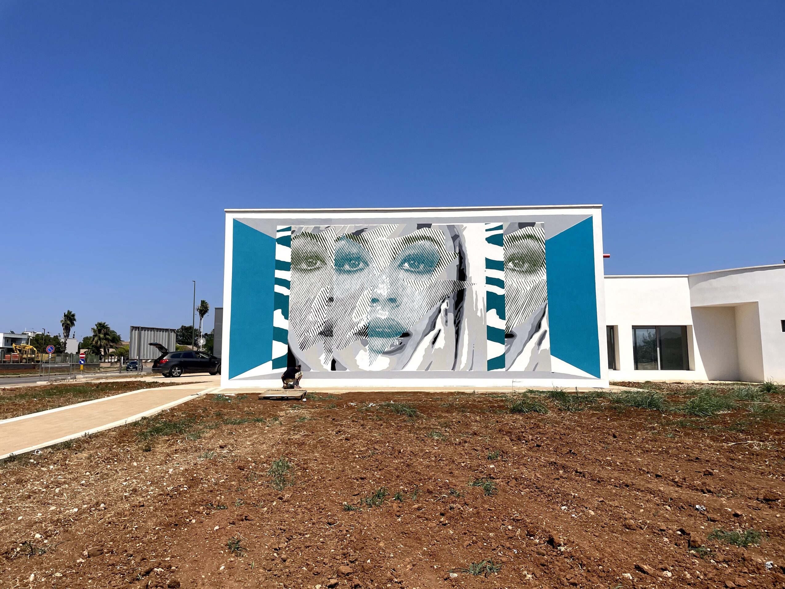 A large mural of a face on a modern white building beside a bare dirt lot under a clear blue sky.