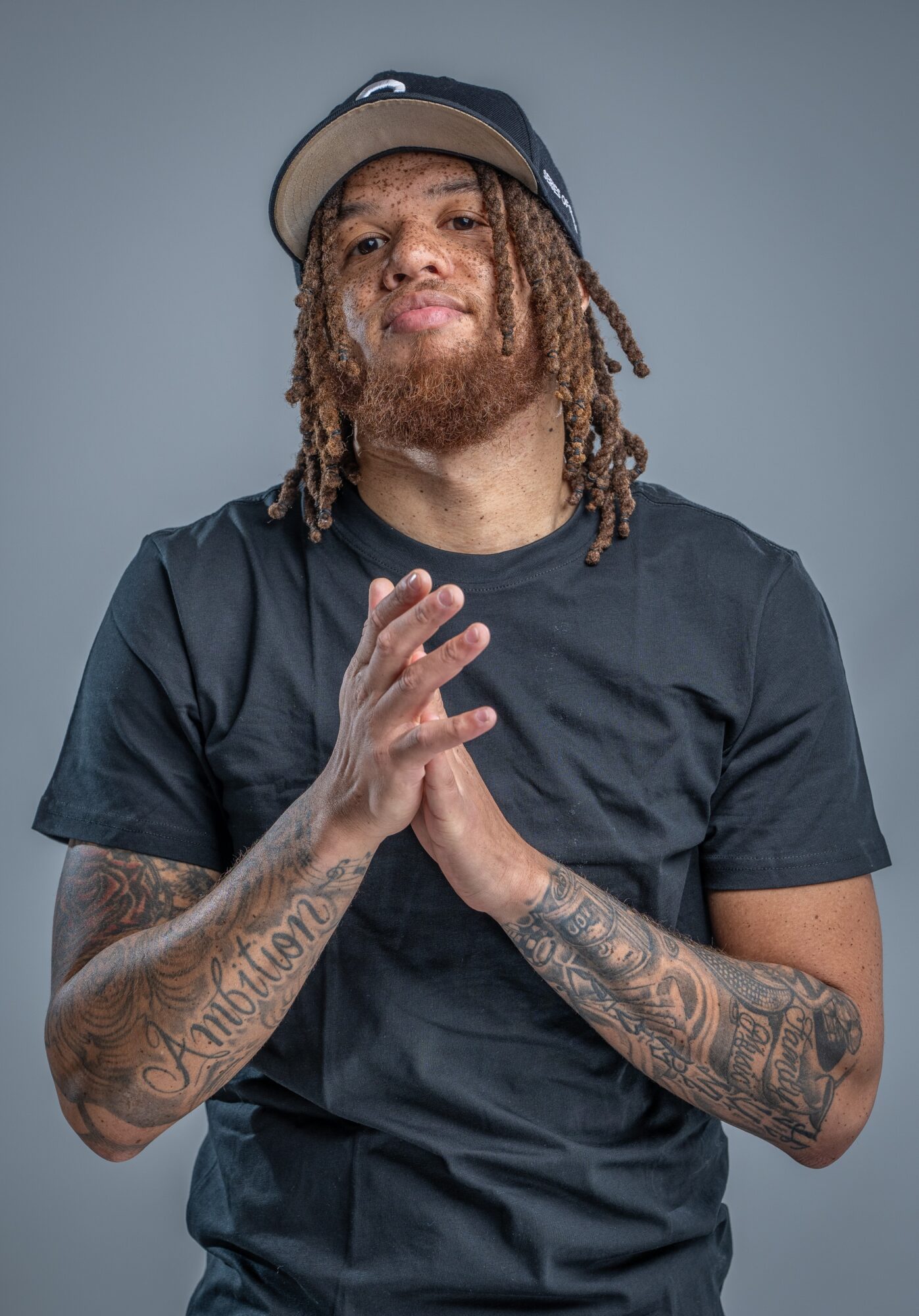 Person wearing a cap and dark shirt, with hands together and tattooed arms, against a gray background.