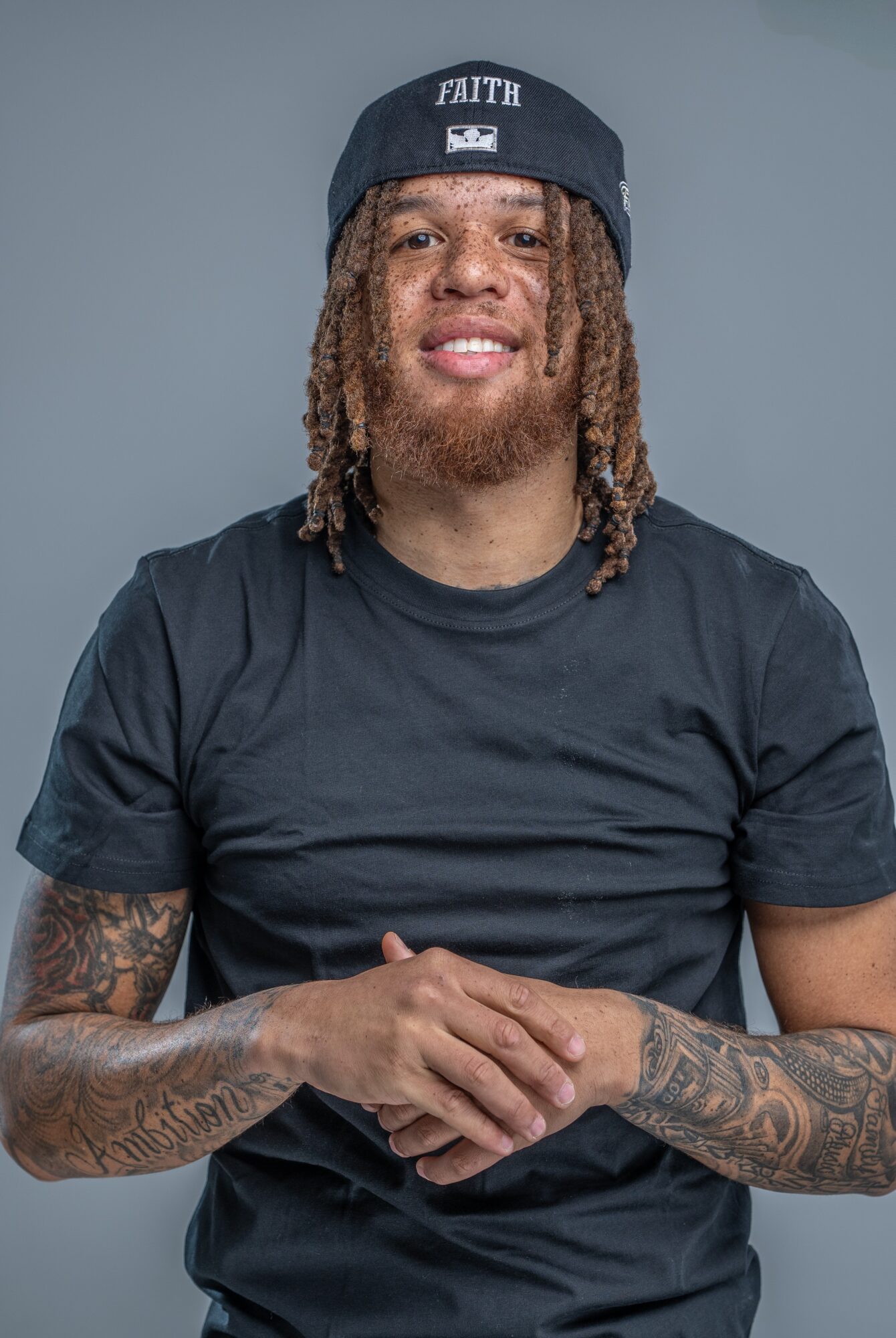 A seated person wearing a black cap and shirt, with tattooed arms against a gray background.