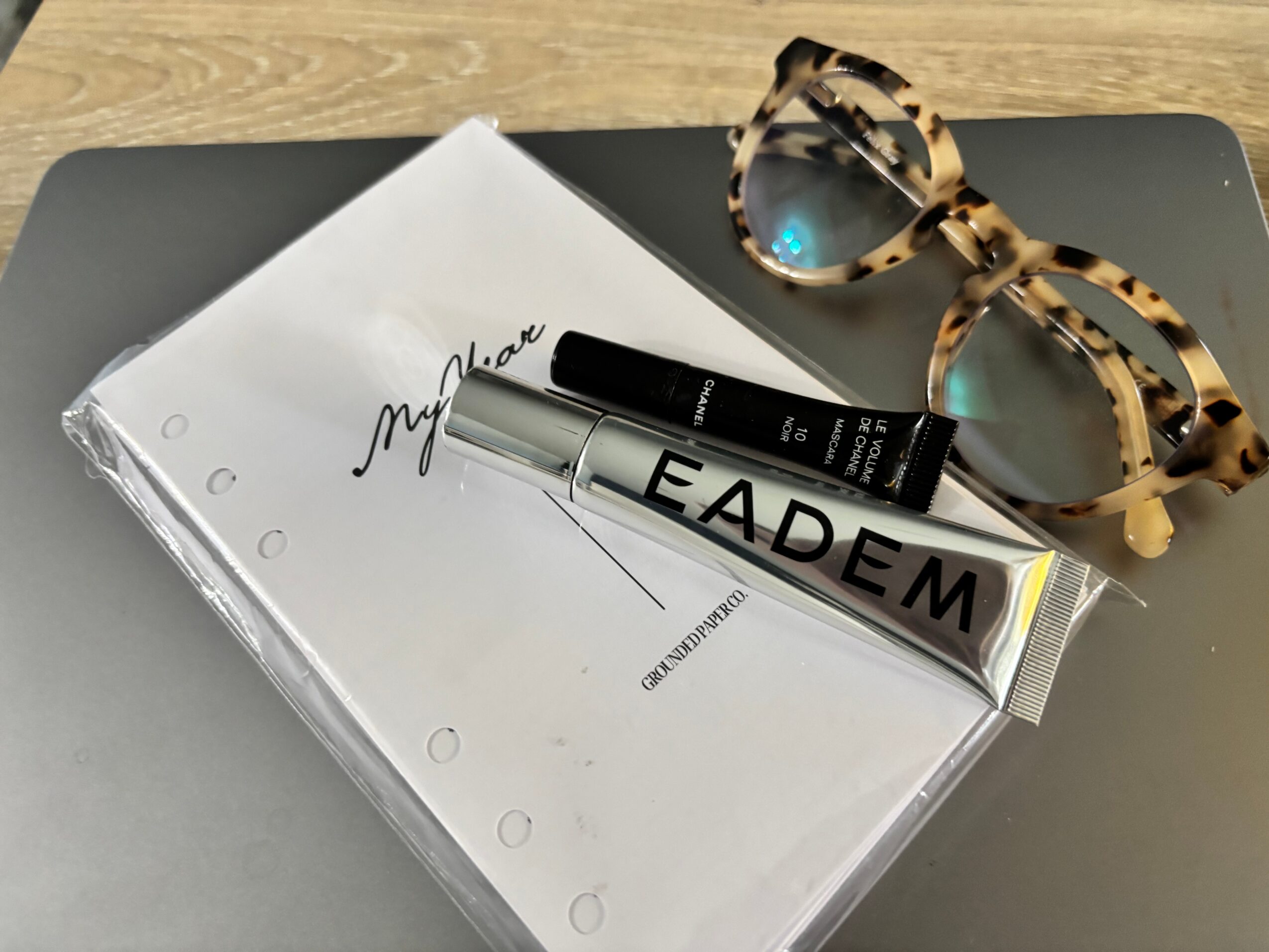A silver cosmetics tube and black pen rest on a white notebook beside tortoiseshell glasses.