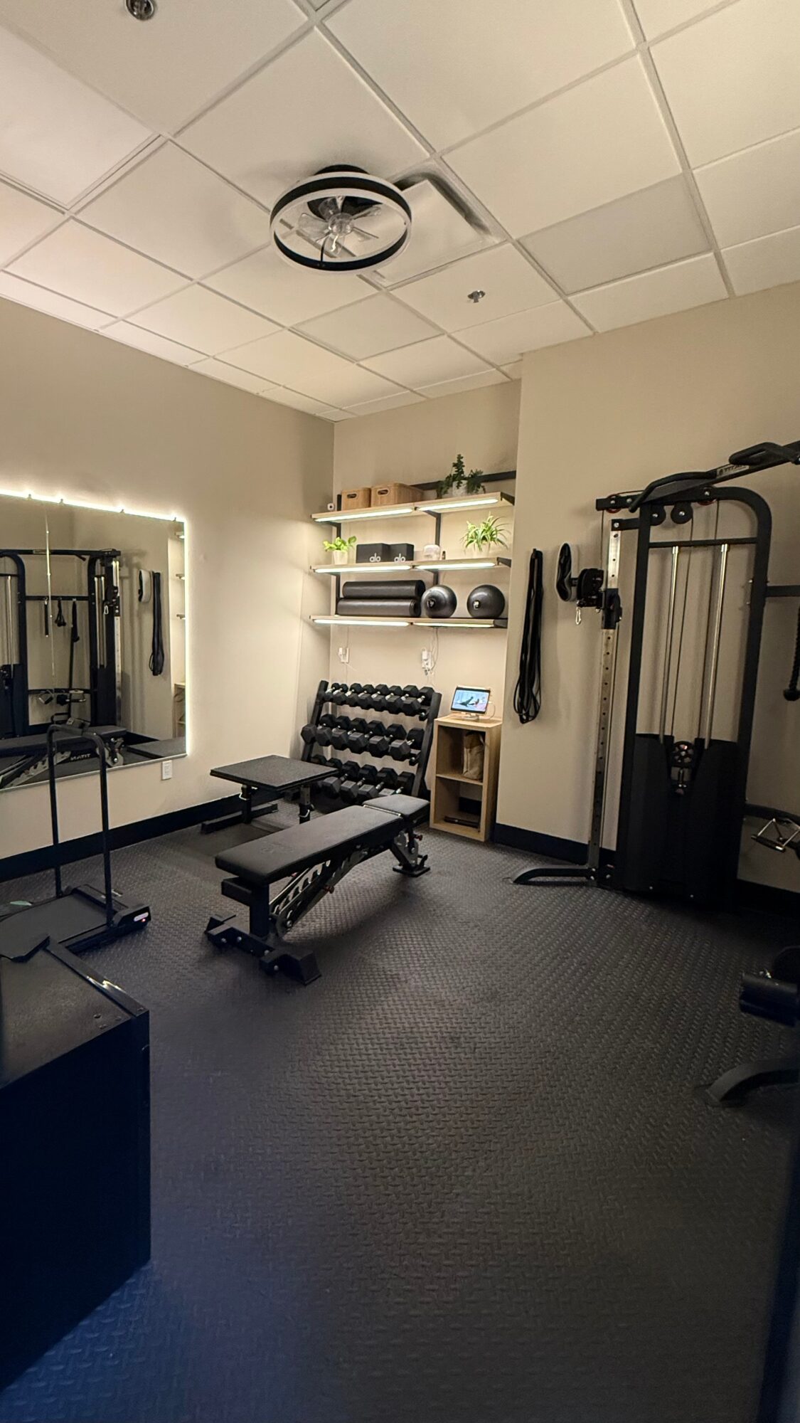 A compact gym with weight benches, dumbbells, cable machines, mirrors, and a tiled ceiling.