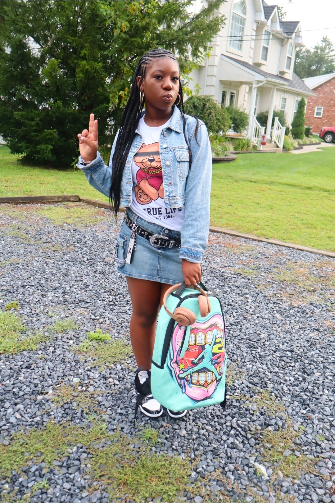 Person standing on a gravel path beside a colorful rolling bag, with houses and trees in the background.