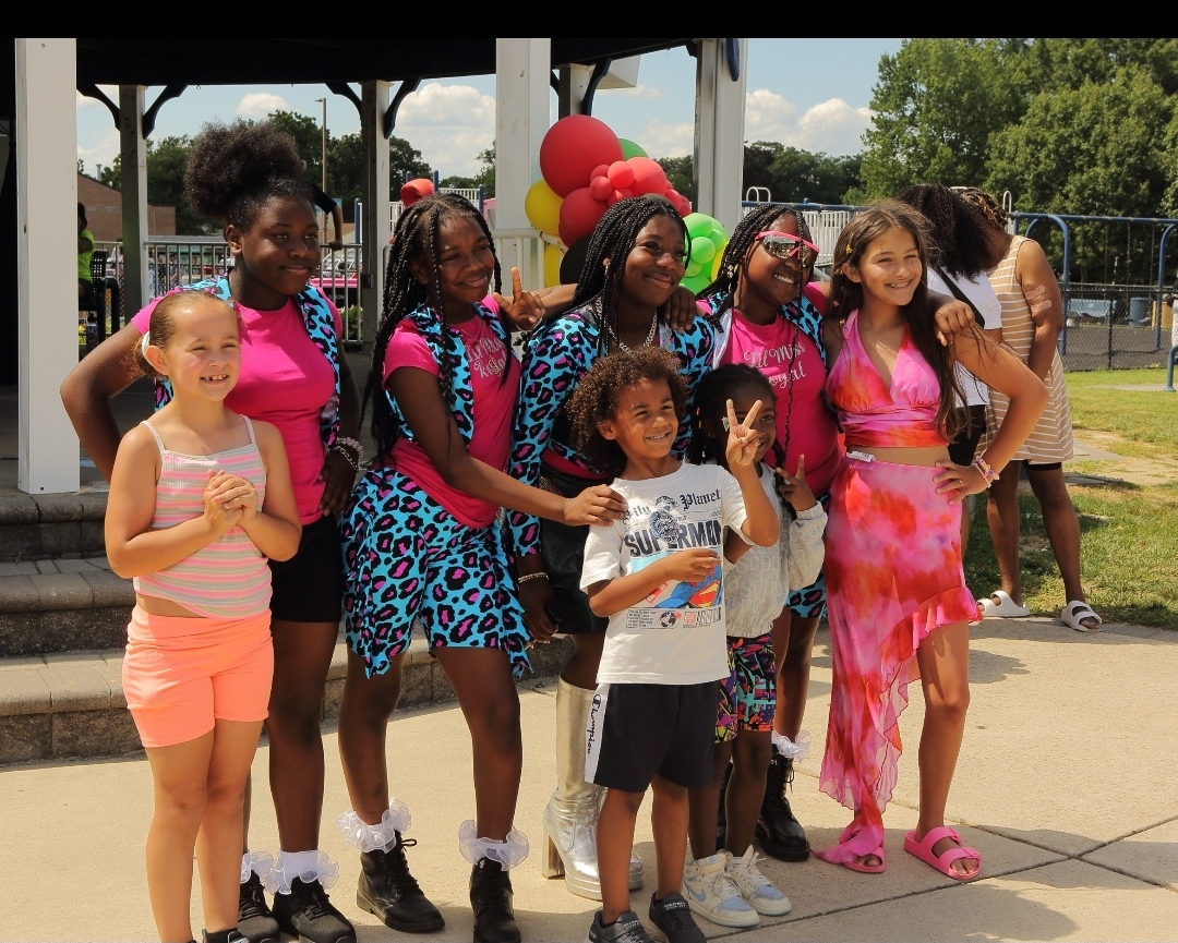 A group of people pose together outdoors beneath a pavilion, with colorful balloons behind them.