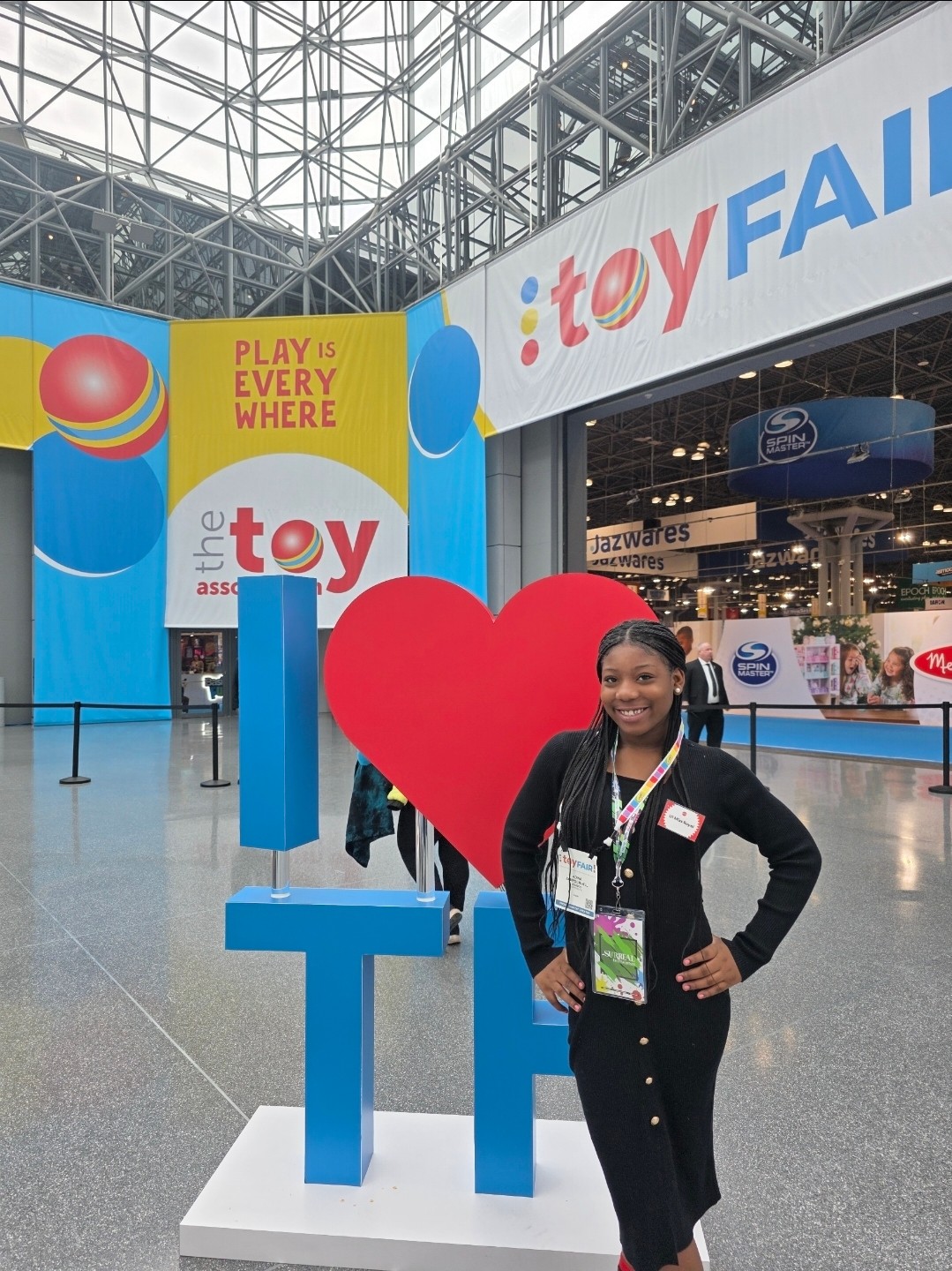 Person standing beside a large red heart and blue display letters at a toy fair booth.