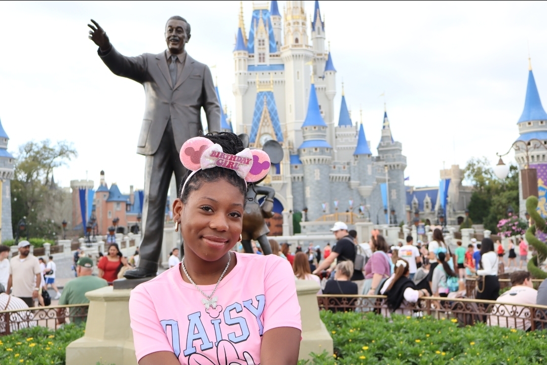 A person wearing pink Minnie Mouse ears poses beside a bronze statue, with a castle and visitors in the background.