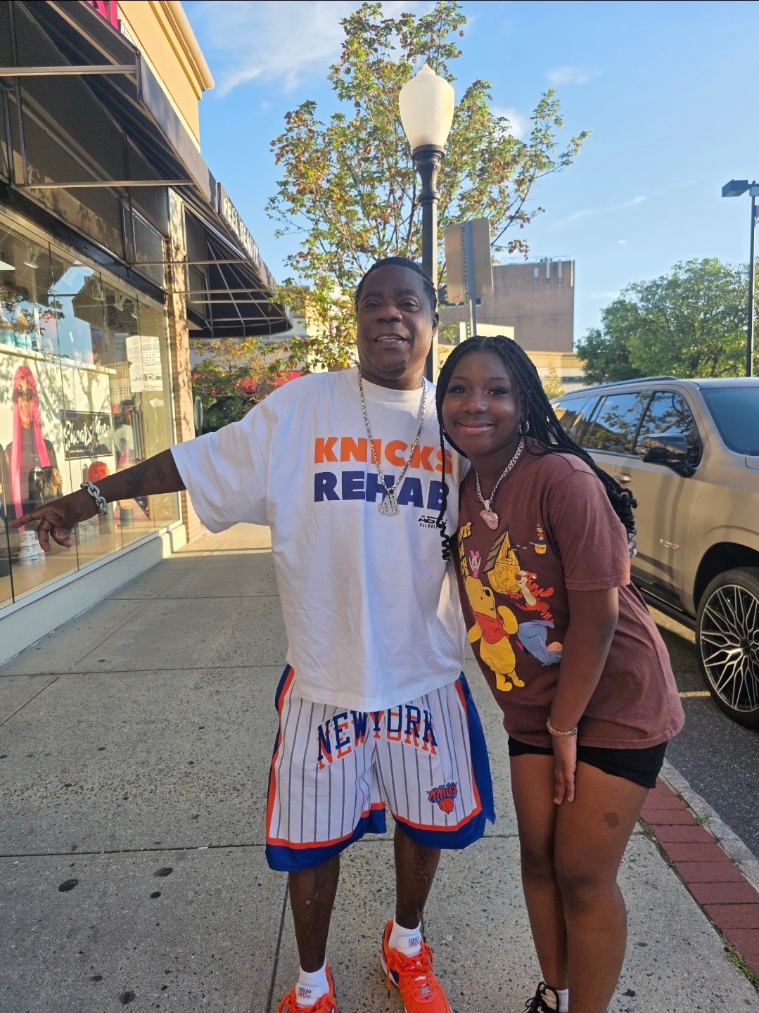 Two people stand on a sidewalk beside a storefront and parked vehicle.