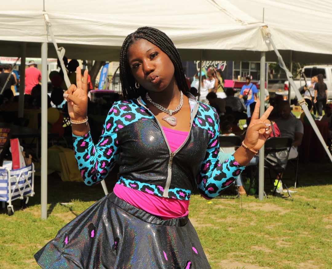 Person in a colorful black outfit posing with both hands raised beneath a canopy at an outdoor event.