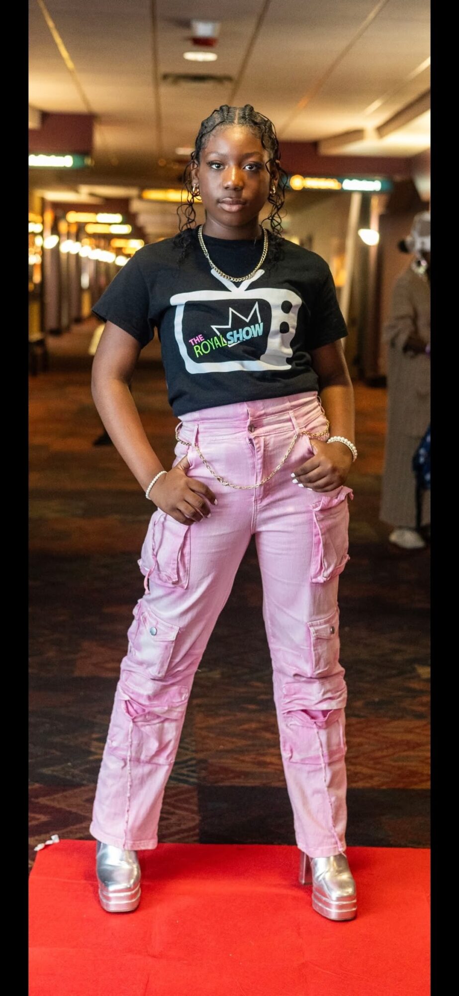 Person wearing a black graphic shirt and pink pants stands on a red carpet indoors.