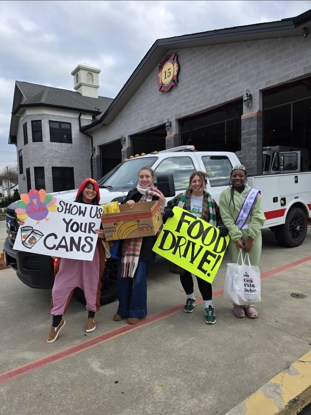 Four people hold food donation signs and boxes in front of a fire truck and building.
