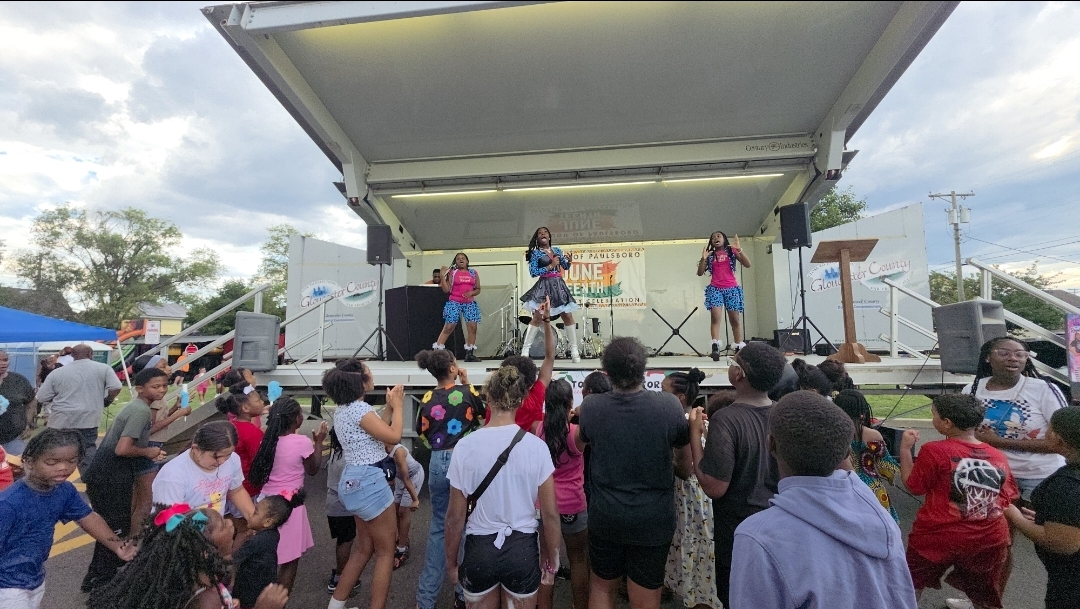 A crowd watches performers on a small outdoor stage beneath a canopy.
