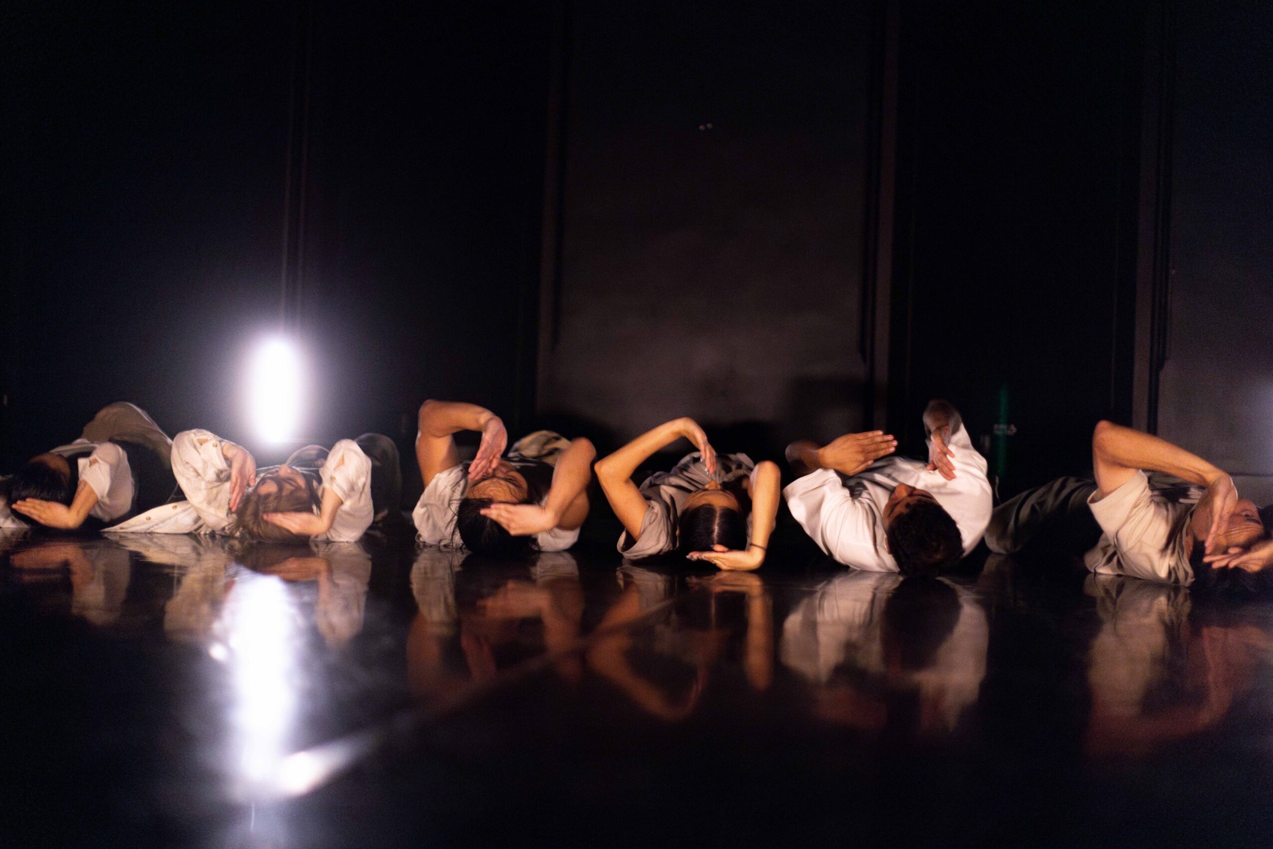 Performers lie in a line on a reflective stage under dim lighting.