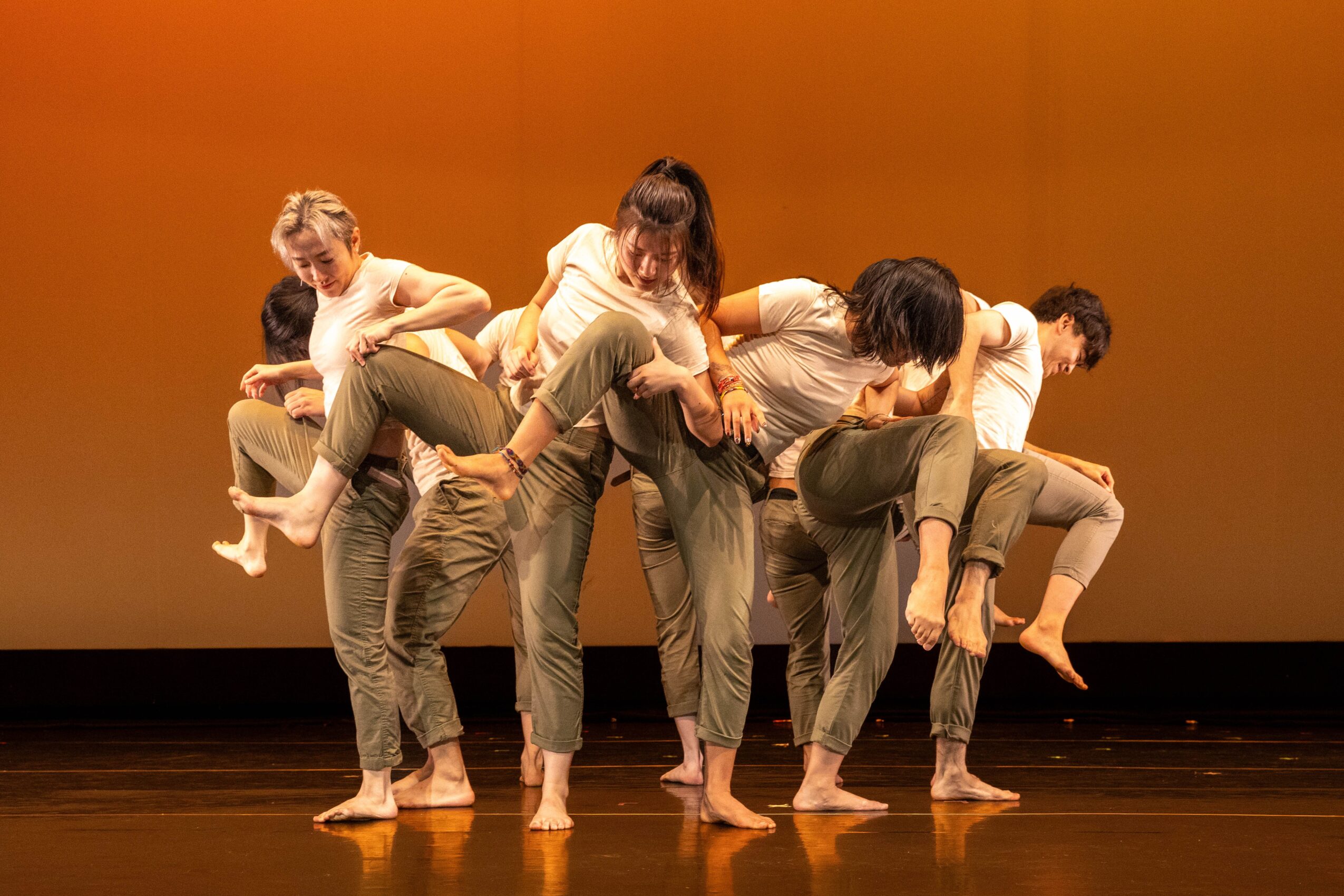 Group of dancers in casual clothing form a layered pose on a stage against an orange backdrop.