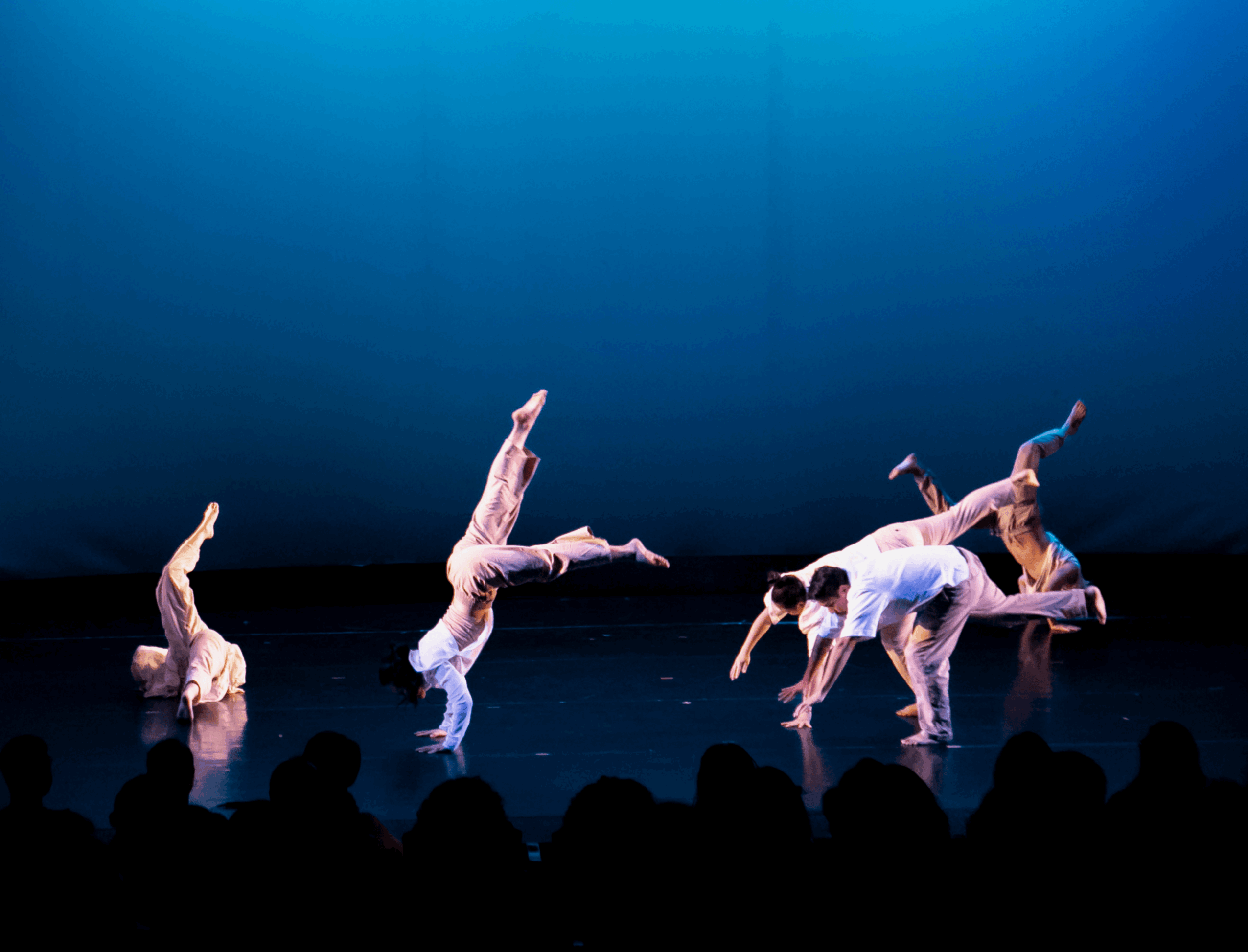 Dancers perform on a stage against a blue backdrop, with seated audience silhouettes in the foreground.